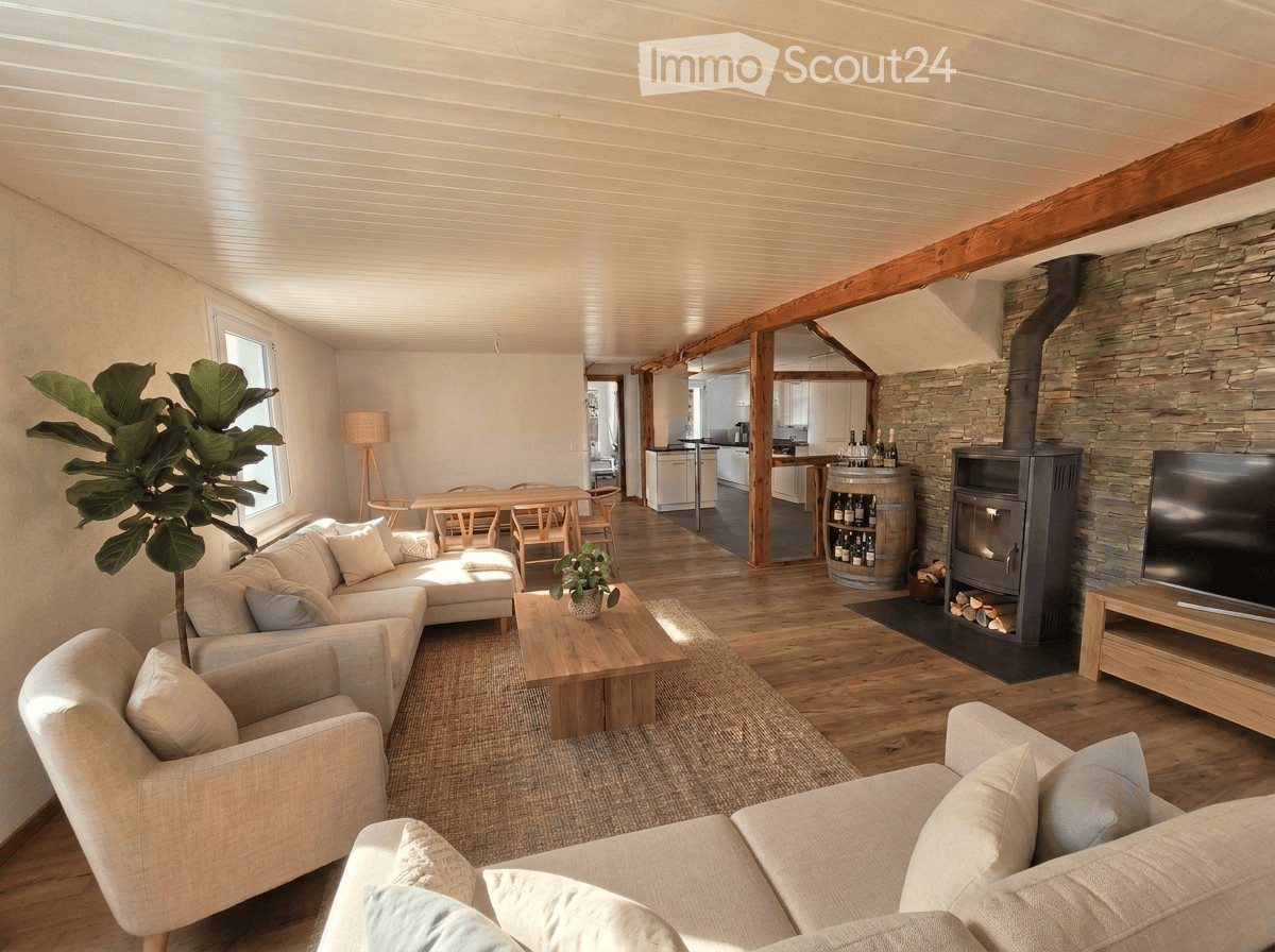Open-plan living and dining area with exposed wooden beams, a stone fireplace, a sofa, coffee table, armchair, dining table with chairs, and a bar area with a barrel and bottles.