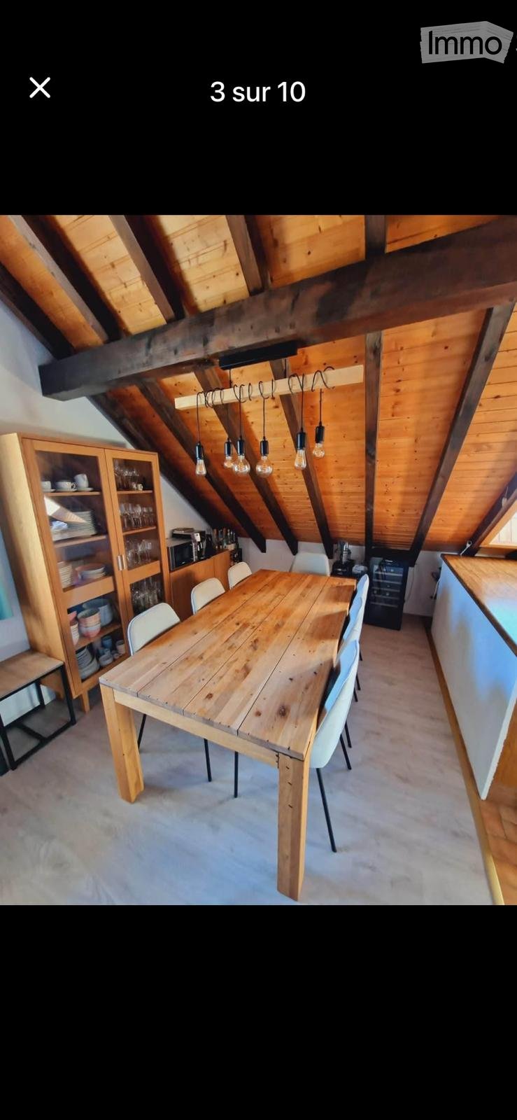 A cozy attic dining area with exposed wooden beams, a large wooden dining table surrounded by white chairs, and built-in wooden cabinets and shelves.
