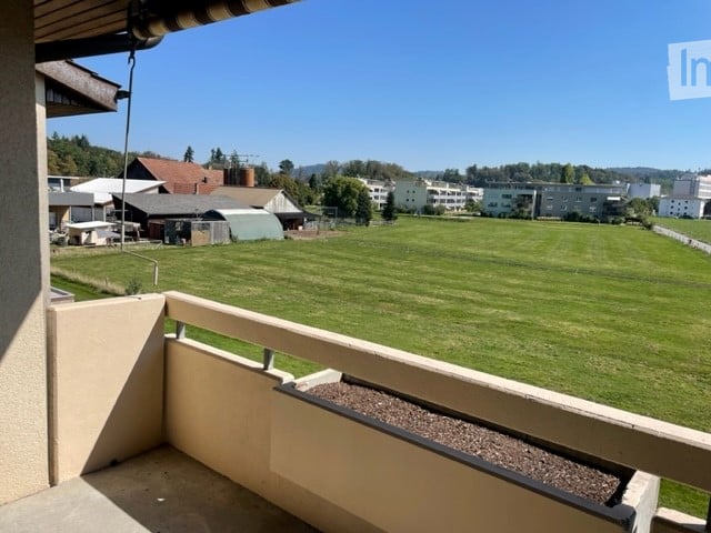 Balcony overlooking a large green field with distant buildings and trees under a clear blue sky.