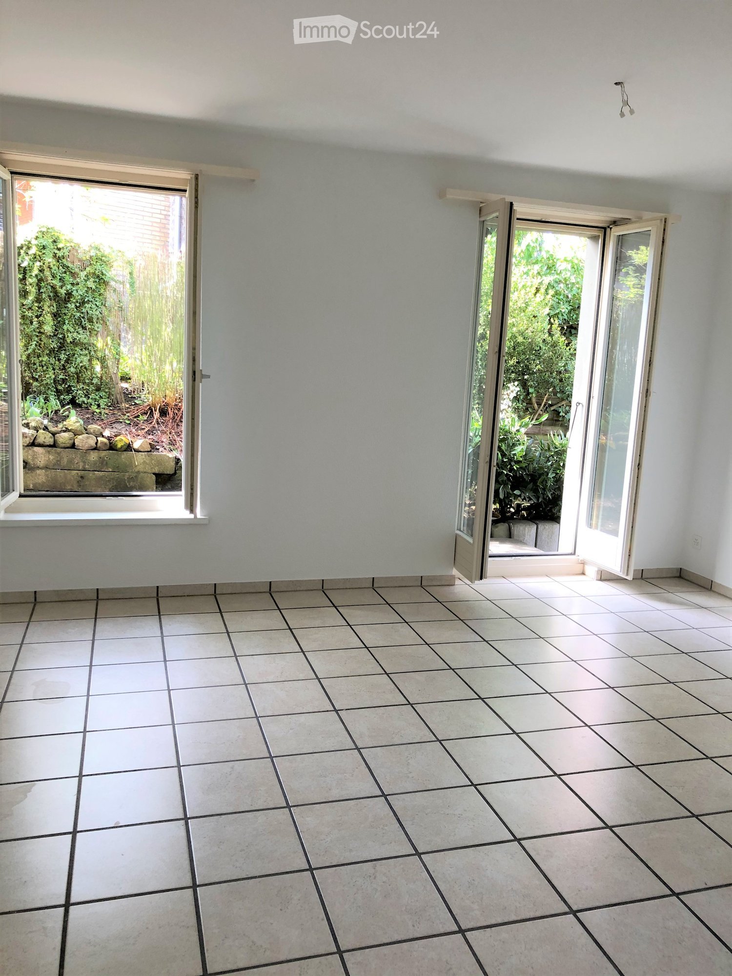 Empty room with white tiled flooring and light-colored walls, featuring two large windows and a sliding glass door leading to a garden area.