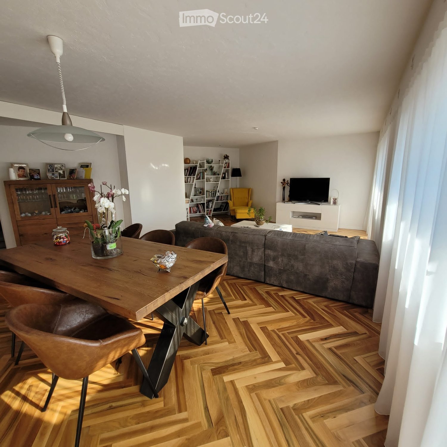Modern living and dining area with wooden herringbone flooring, grey sectional sofa, wooden dining table with four chairs, television on a white stand, bookshelf, and decorative cabinet.