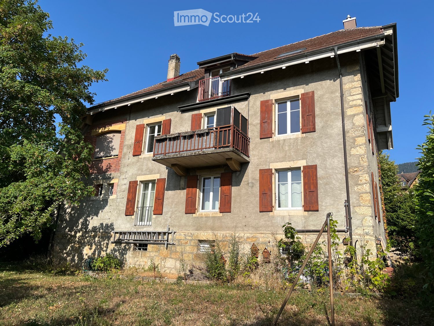 Two-story stone house with multiple windows, wooden shutters, and a balcony on the upper floor.
