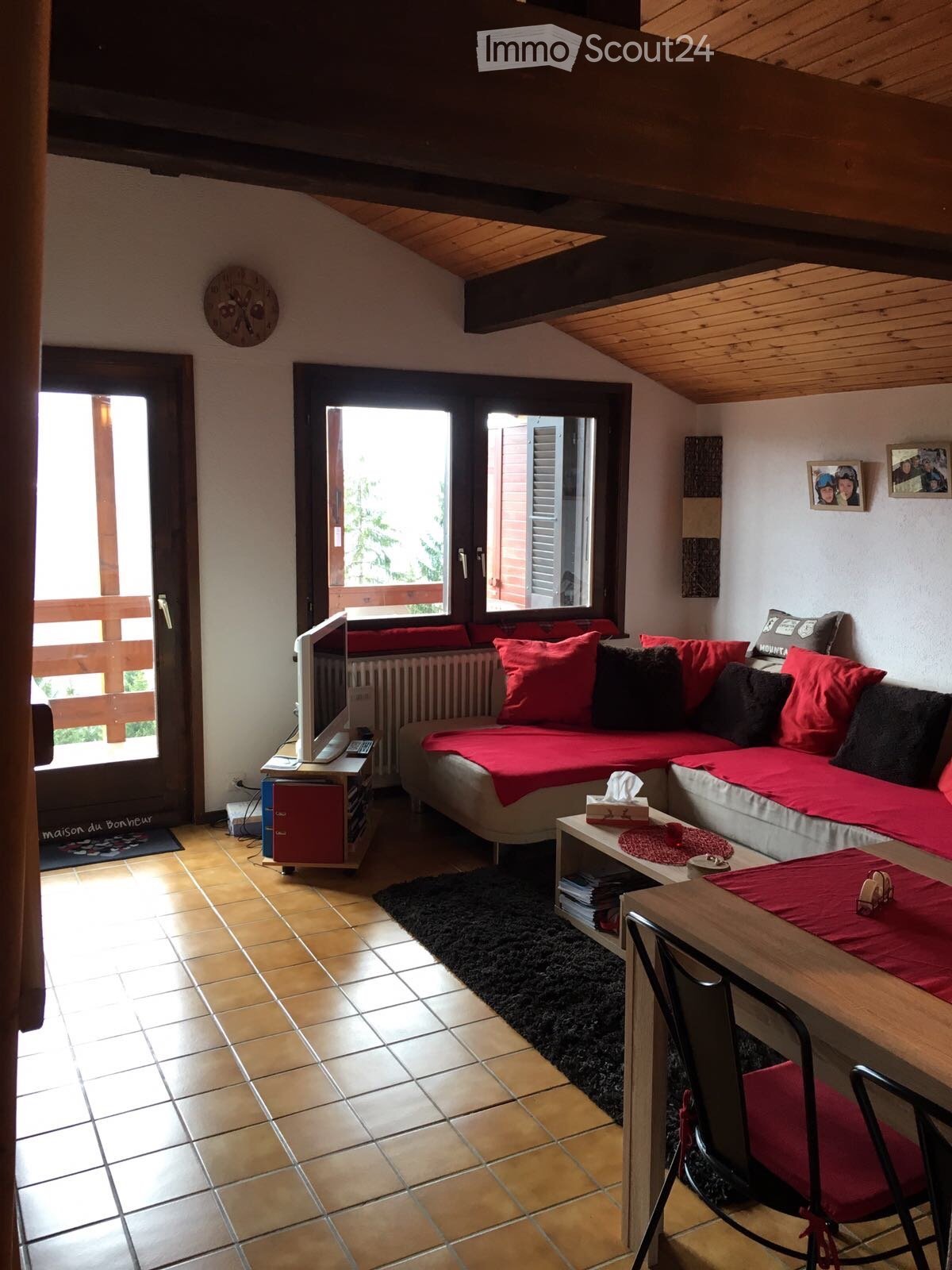 Cozy living room with a sectional sofa, coffee table, TV, and a dining area with a table and chairs. The room features wooden beams on the ceiling and large windows with a balcony view.