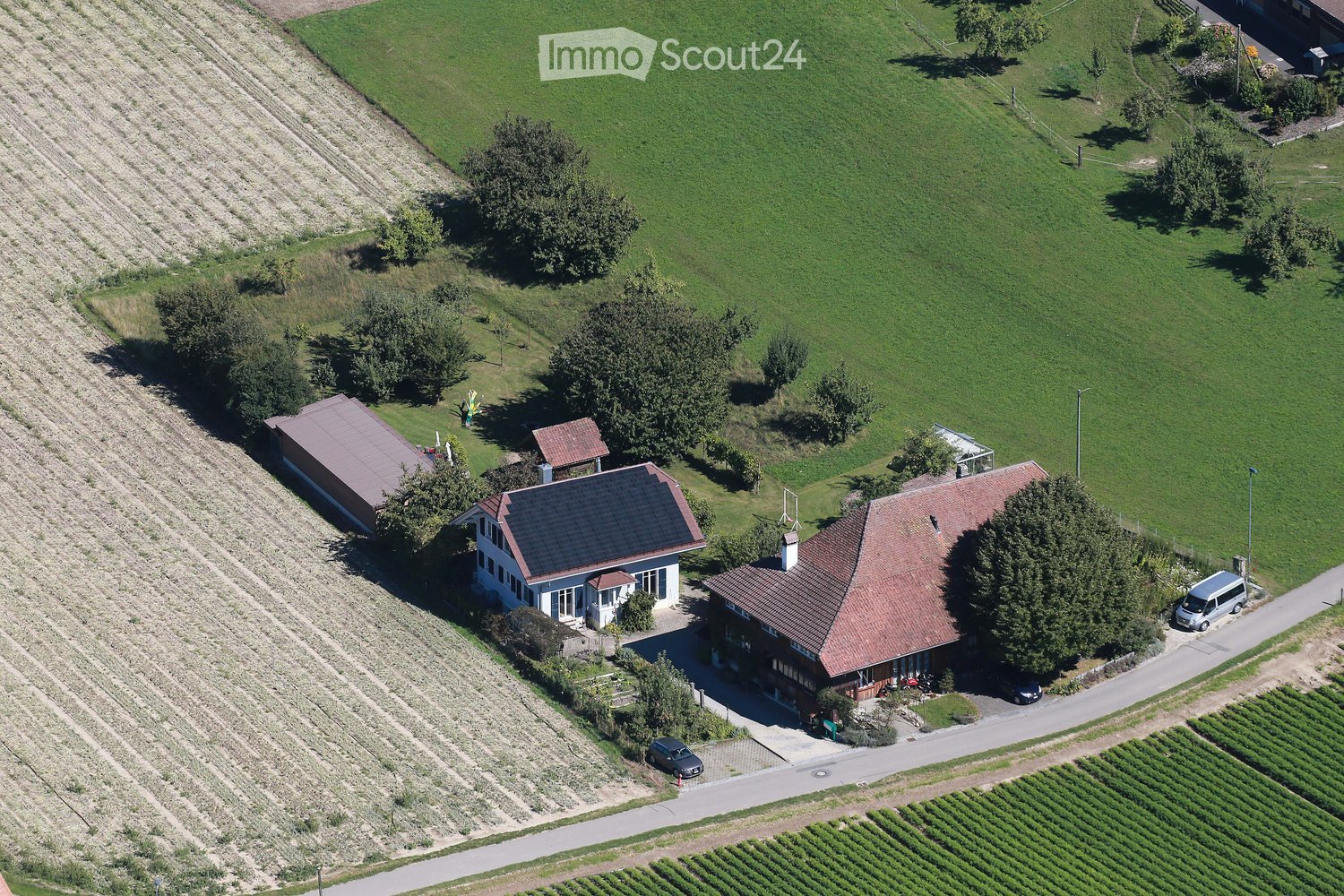 Aerial view of a residential property with multiple buildings, including a main house, a garage, and outbuildings, surrounded by green fields and vineyards.