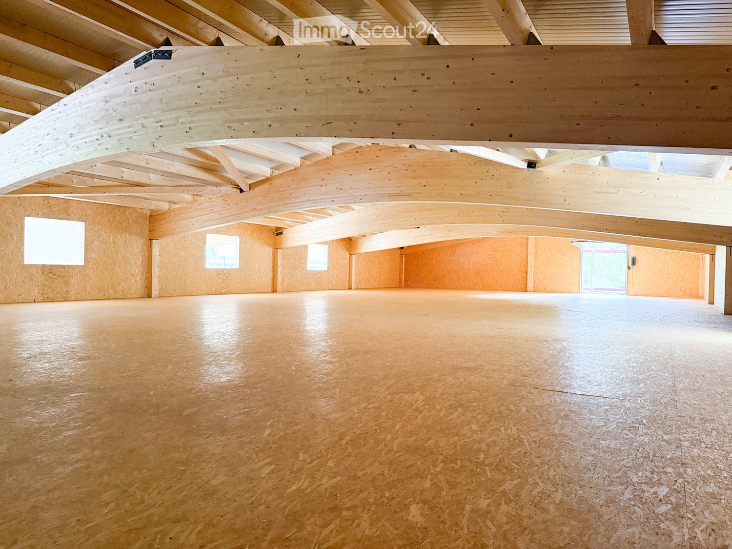 Empty room with wooden floor and exposed wooden beams on the ceiling. Walls are covered in light wood paneling, with several windows allowing natural light to enter.
