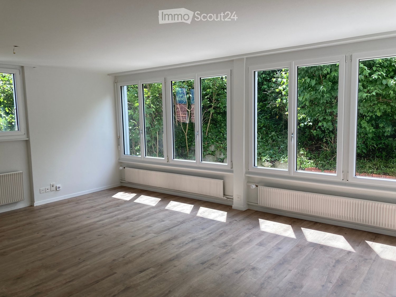 Empty room with wooden flooring, large windows, radiators, and white walls.