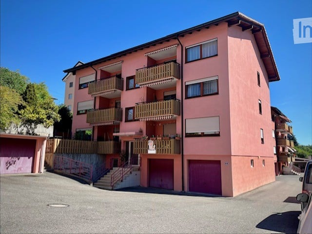 A three-story pink building with multiple balconies and a staircase leading to the entrance. The building features several garage doors and is situated in a residential area with a clear blue sky in the background.