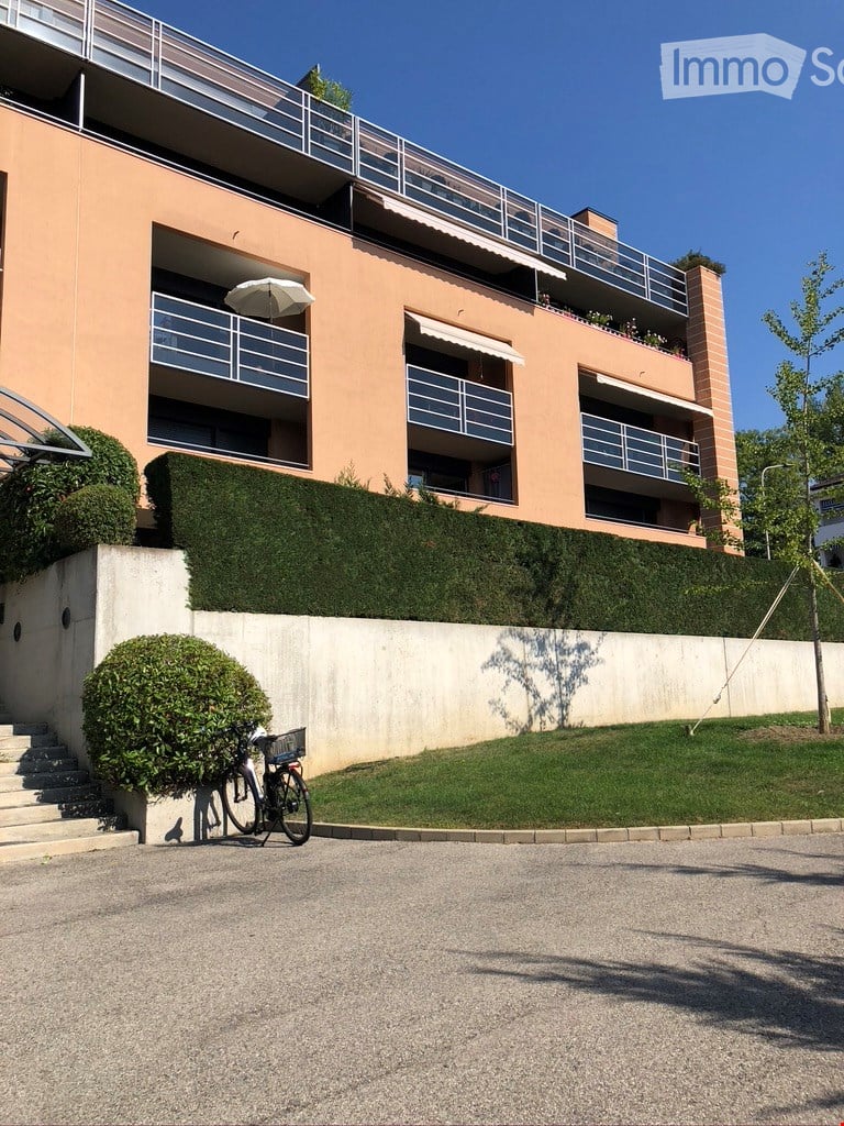 Modern multi-story building with balconies, light pink facade, and neatly trimmed hedges. A bicycle is parked near the entrance.