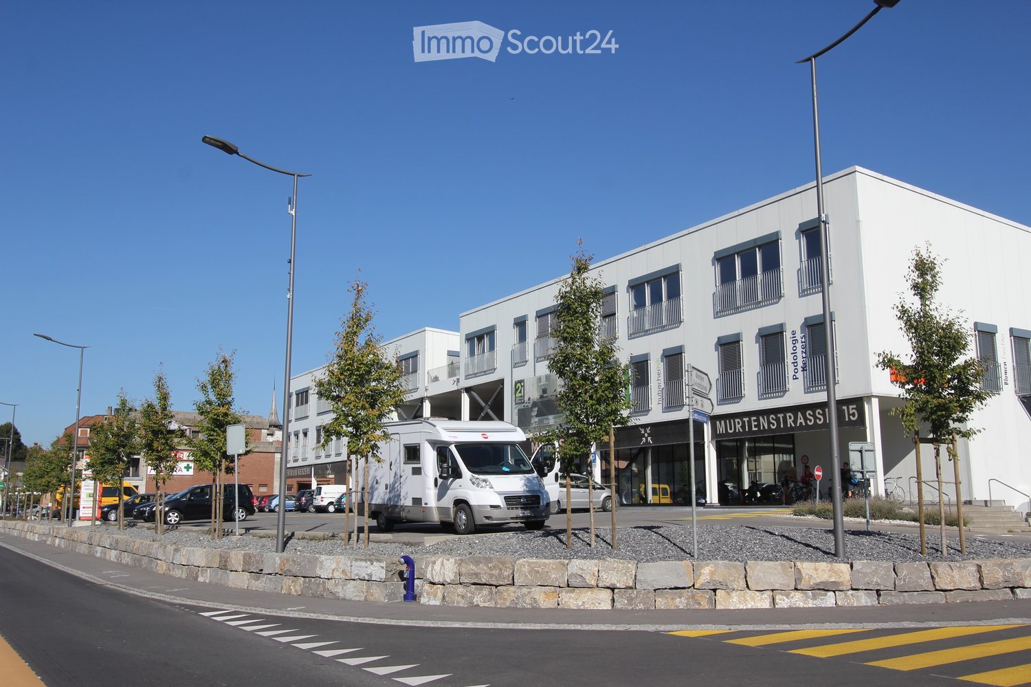 Modern multi-story building with white facade, large windows, and balconies. A camper van is parked in front, and there are several cars and streetlights visible.