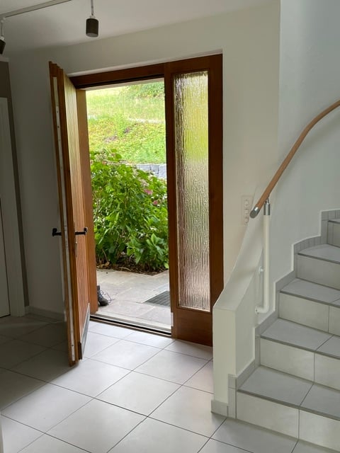 Hallway with tiled floor, open door leading to a garden area, staircase with wooden handrail visible.