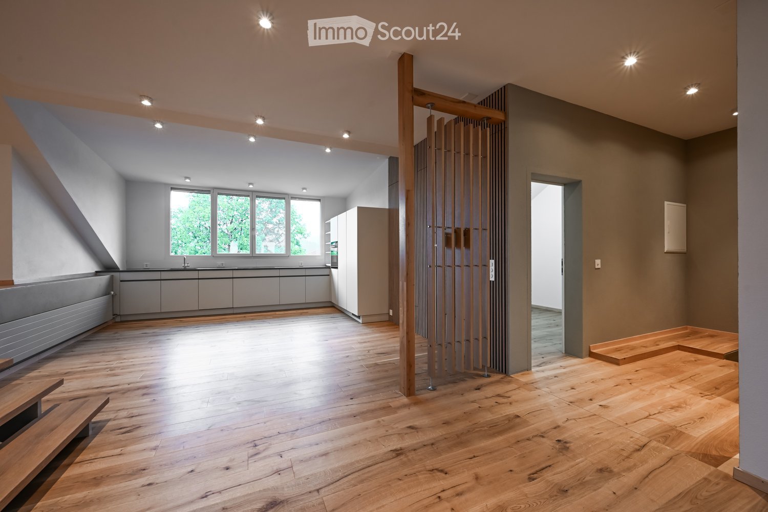 Open-plan living and kitchen area with wooden flooring, modern kitchen cabinets, and a sleek sink. A wooden partition separates the spaces, and recessed lighting illuminates the area.