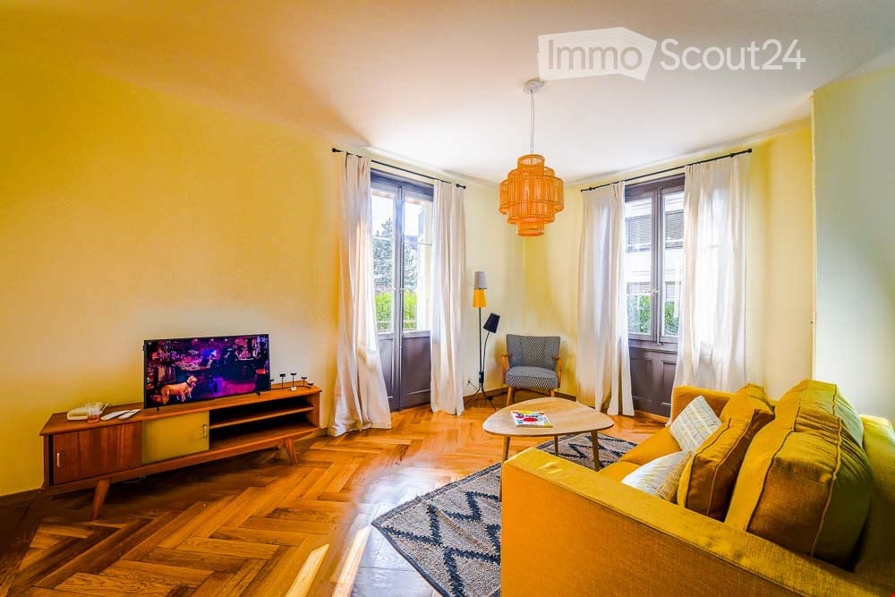 Bright living room with wooden flooring, large windows with white curtains, a yellow sofa, a coffee table, a television on a wooden stand, and a patterned rug.