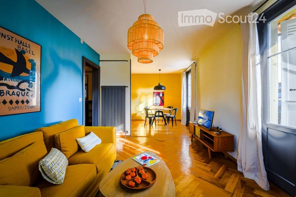 Bright, open-plan living and dining area with wooden flooring, yellow sofa, coffee table with fruit, dining table with chairs, and a television on a wooden stand.