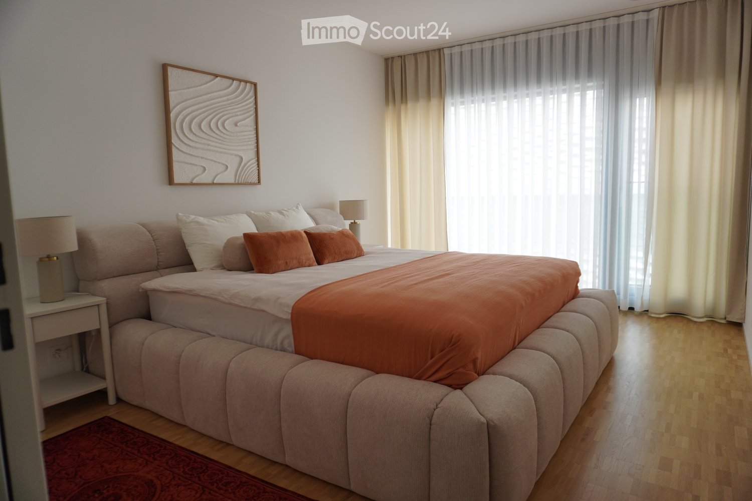 Modern bedroom with a large bed, beige sofa, nightstand with lamp, and a decorative picture frame on the wall.