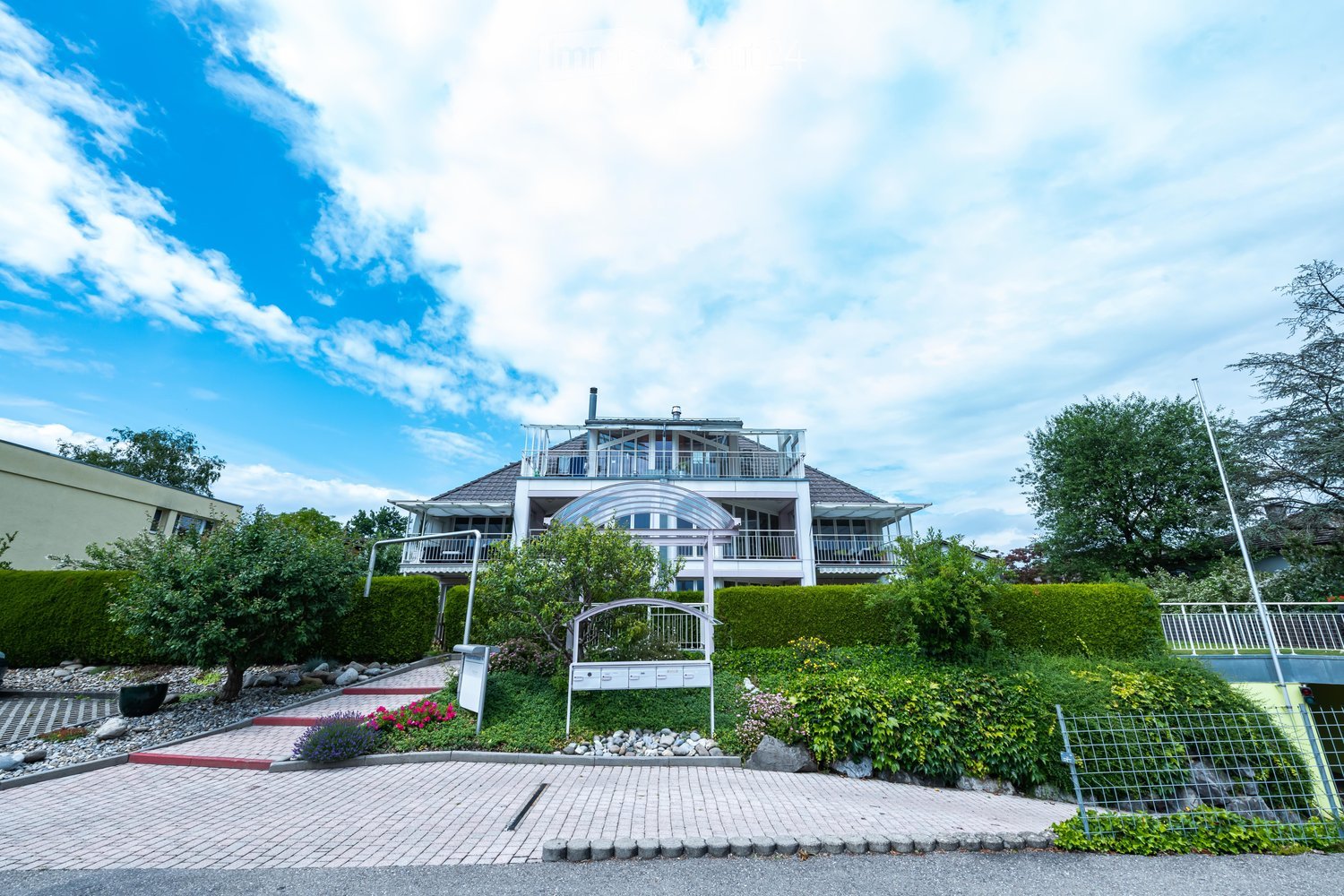 A modern two-story house with a white exterior, large windows, and a balcony on the upper level. The house is surrounded by well-maintained gardens, hedges, and a brick pathway leading to the entrance.