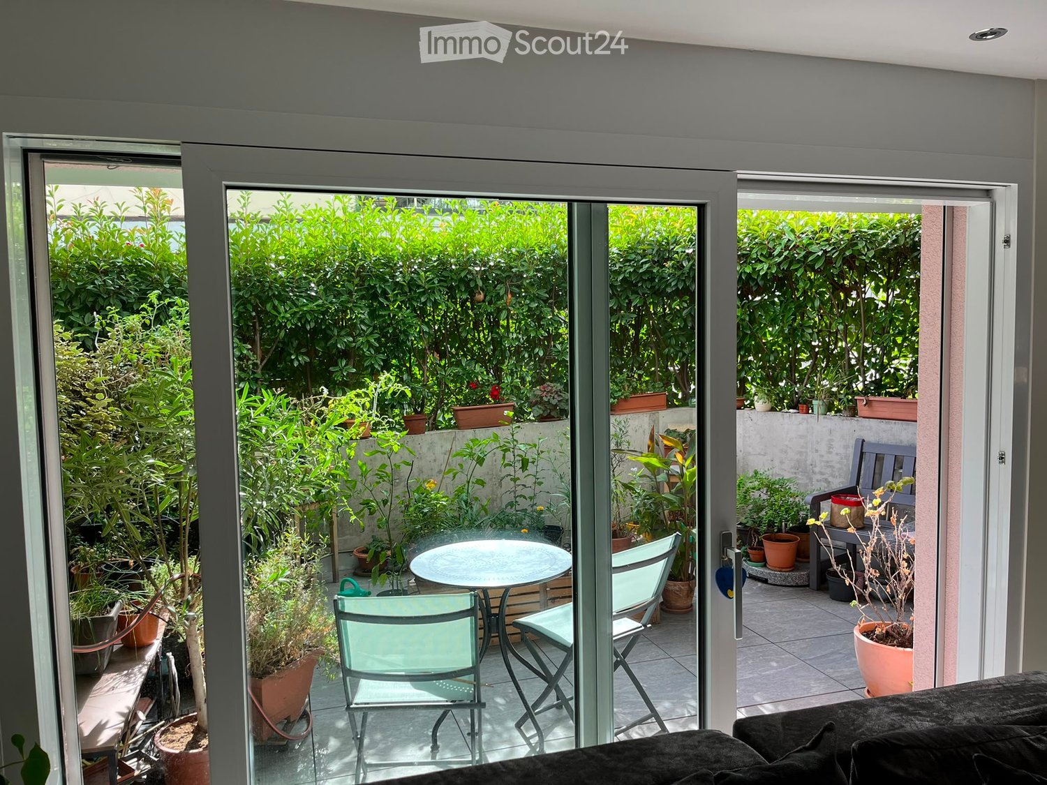 Balcony with glass doors leading to a lush garden, featuring a small table, chairs, a bench, and several potted plants.
