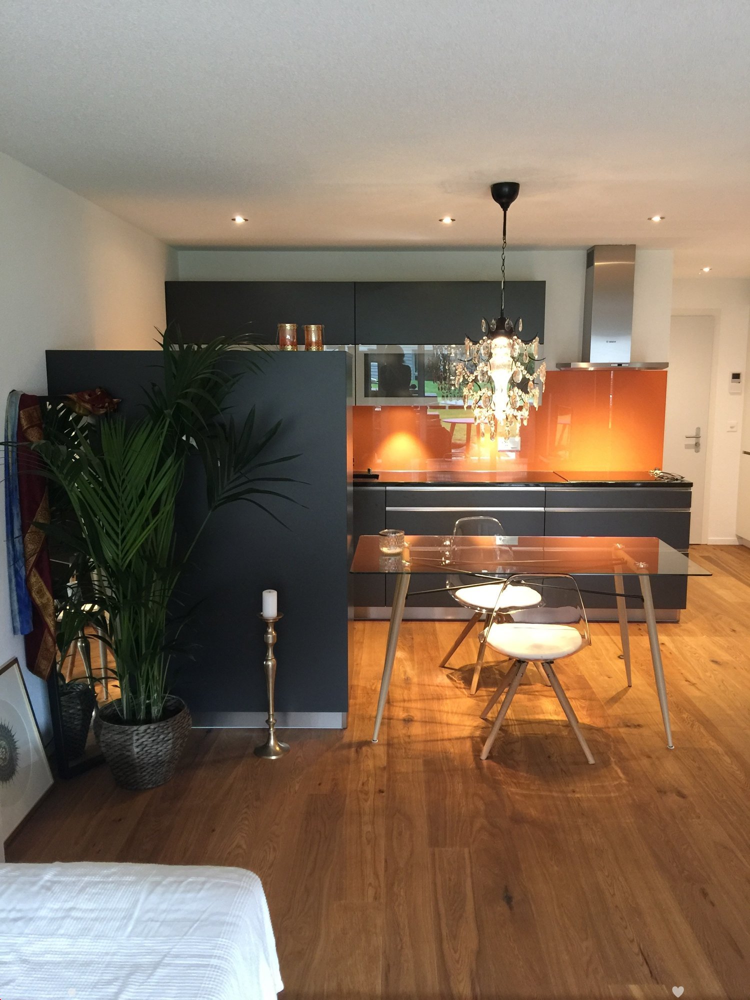 Modern kitchen and dining area with dark grey cabinets, a glass dining table, white chairs, a chandelier, and a potted plant. The floor is wooden, and the kitchen features a stainless steel range hood and a backsplash with an orange accent.