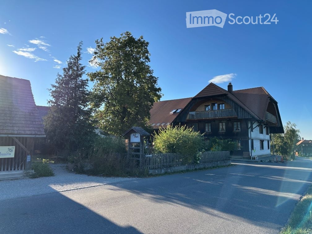 Traditional two-story house with a wooden balcony, surrounded by trees and a wooden fence.