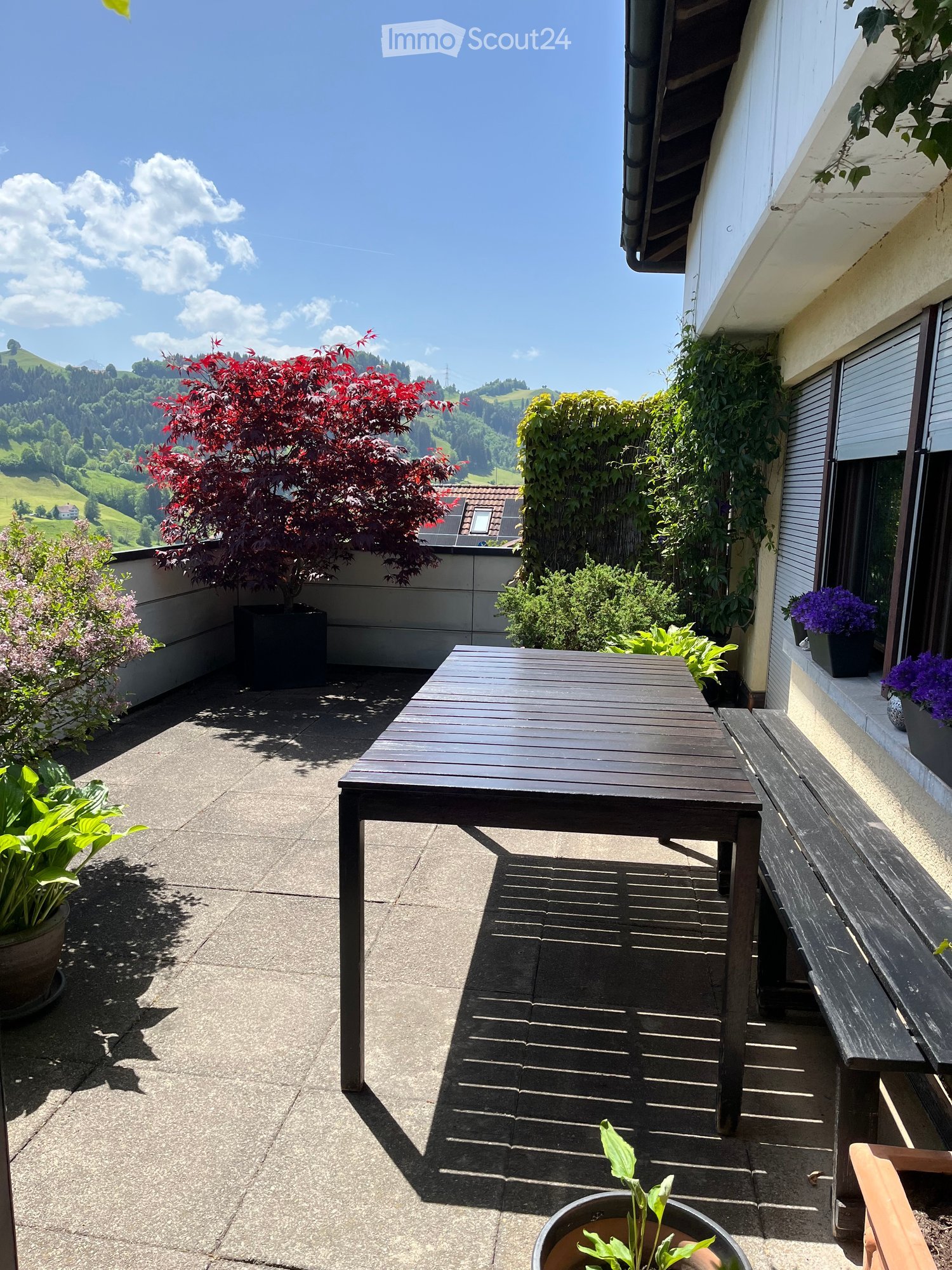 Balcony with a wooden table, bench, and potted plants, overlooking a scenic valley.