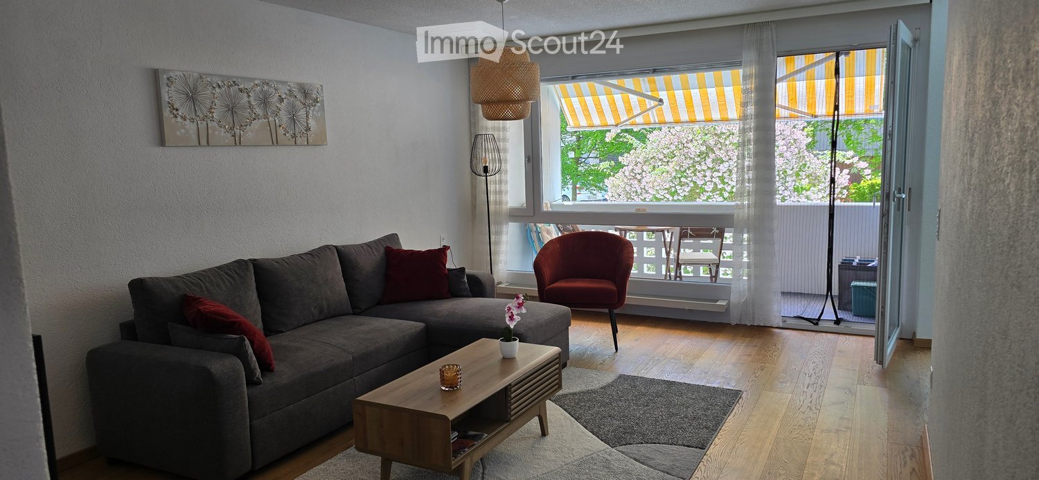 Modern living room with a grey sectional sofa, wooden coffee table, red armchair, and large windows opening to a balcony. Features include a floor lamp, table lamp, and decorative artwork.