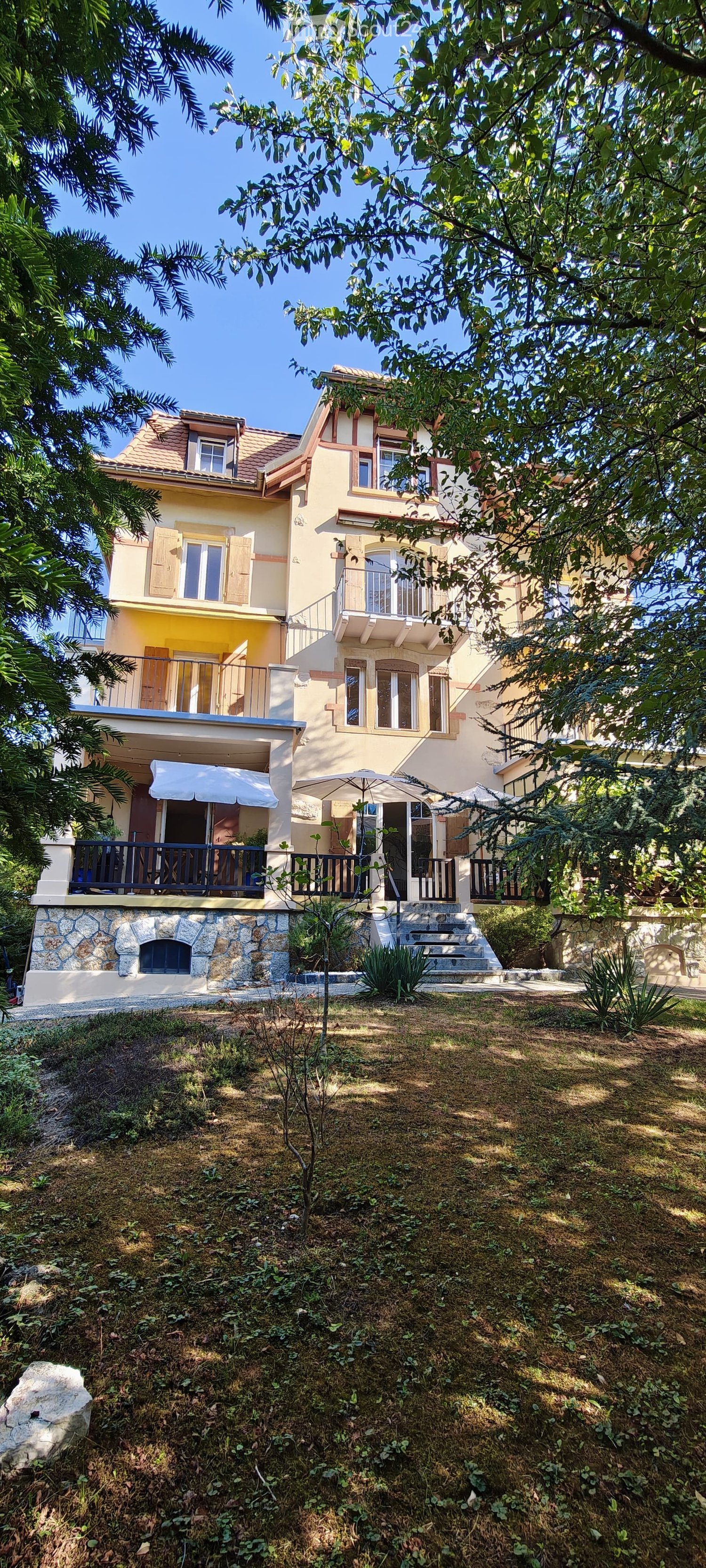 Two-story house with yellow and white exterior, multiple balconies, stone base, and lush greenery surrounding the property.
