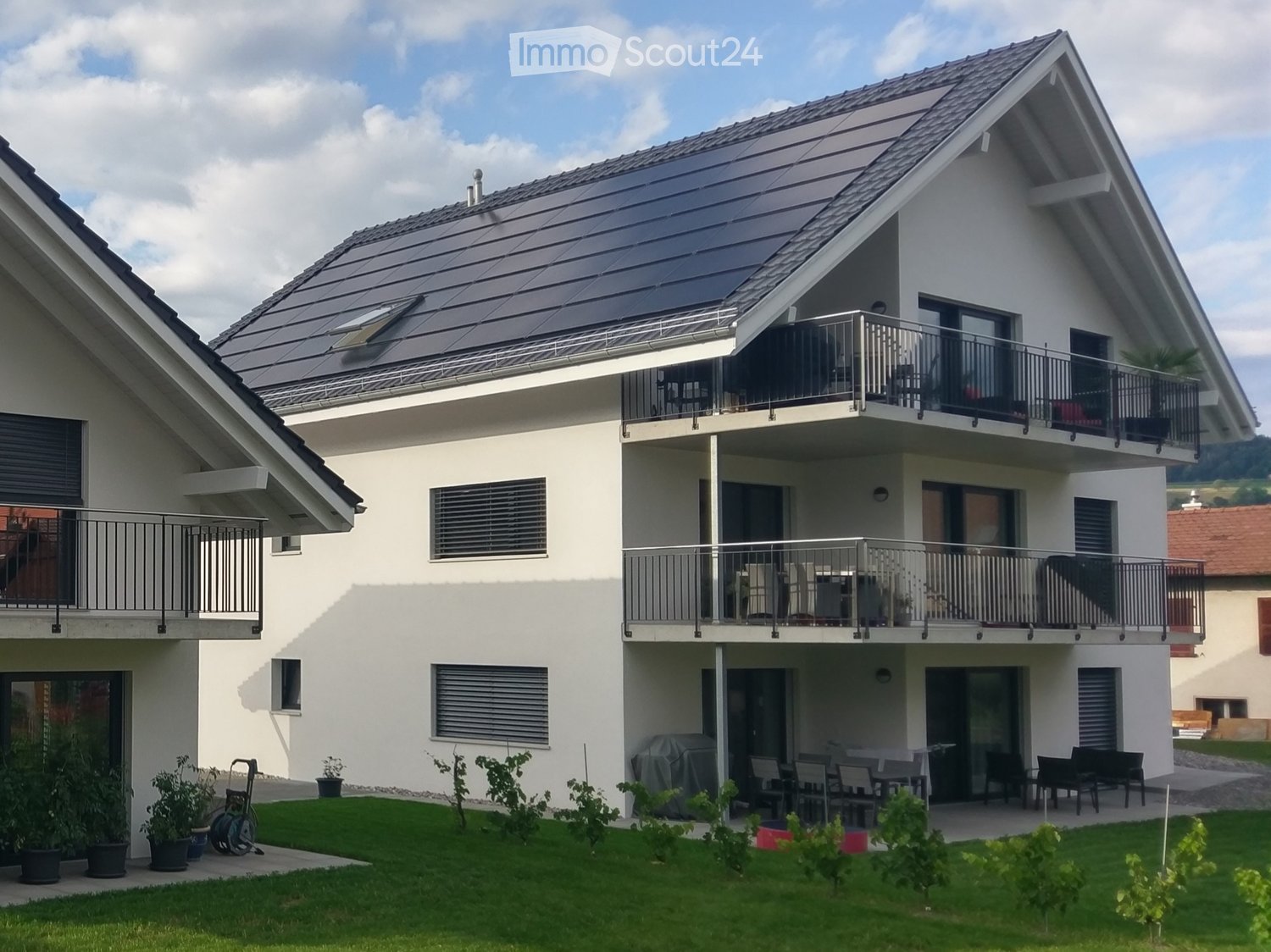 Two-story house with white exterior, solar panels on the roof, multiple balconies, and a well-maintained garden with potted plants and outdoor furniture.