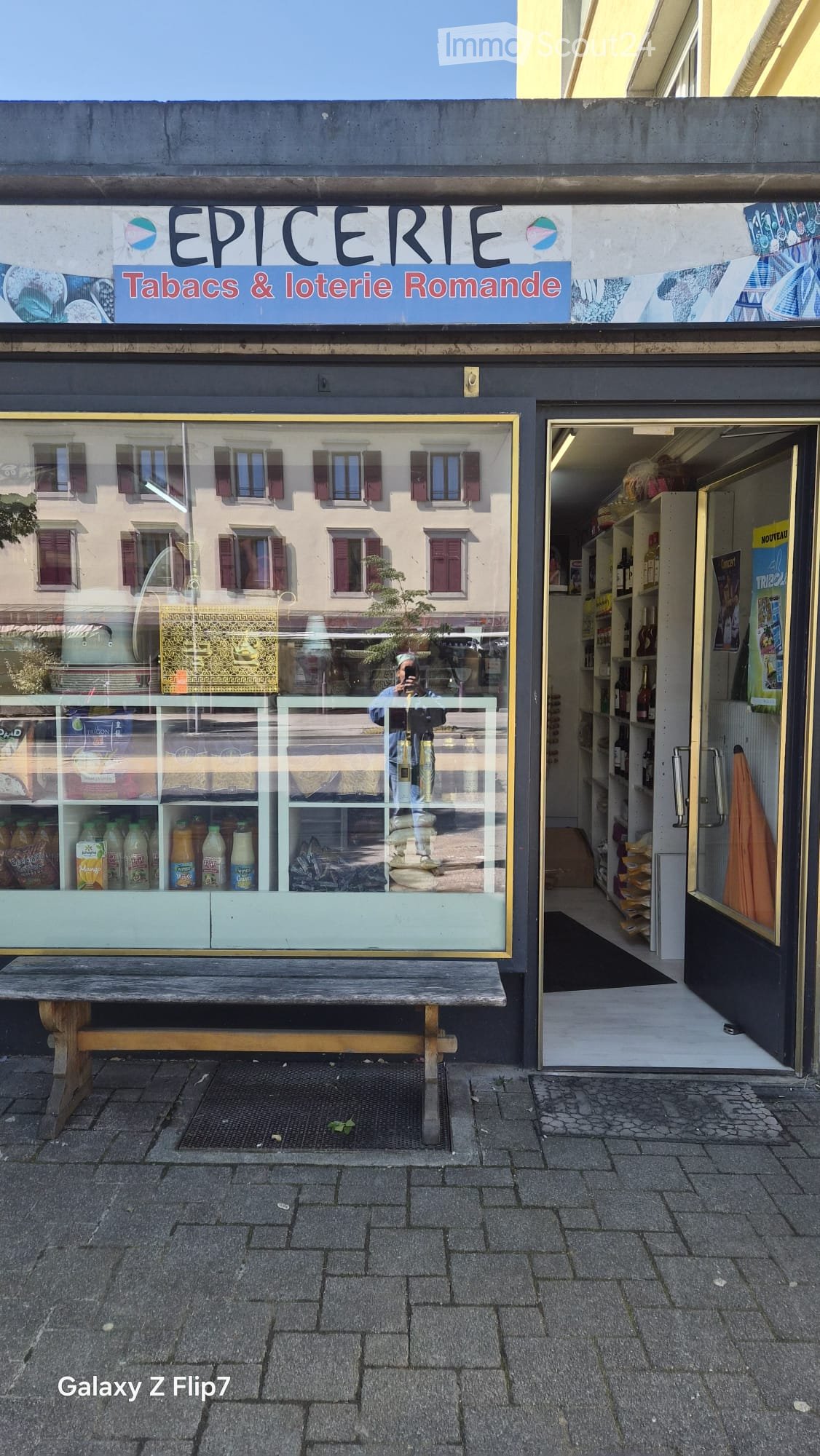 Facade of a small shop named "Epicerie Tabacs & loterie Romande" with a glass display case showing beverages and bread, a wooden bench in front, and a reflection of a building with red shutters in the glass.