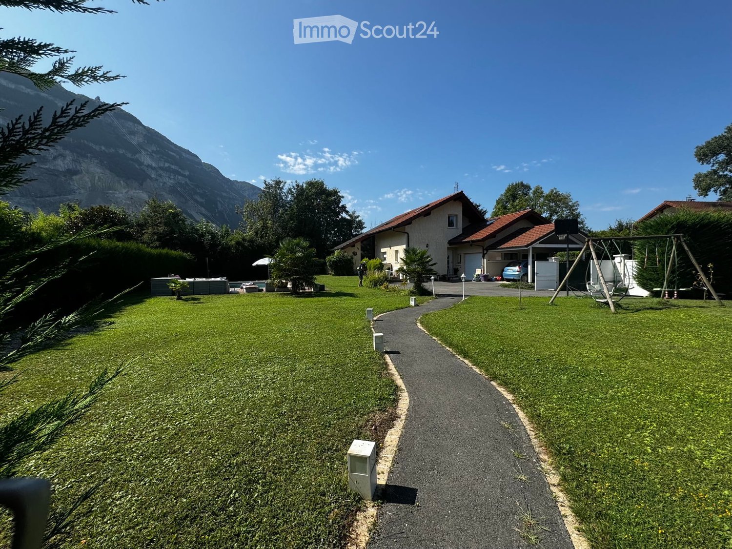 Lush green lawn with a curved pathway leading to a house, surrounded by trees and mountains in the background.