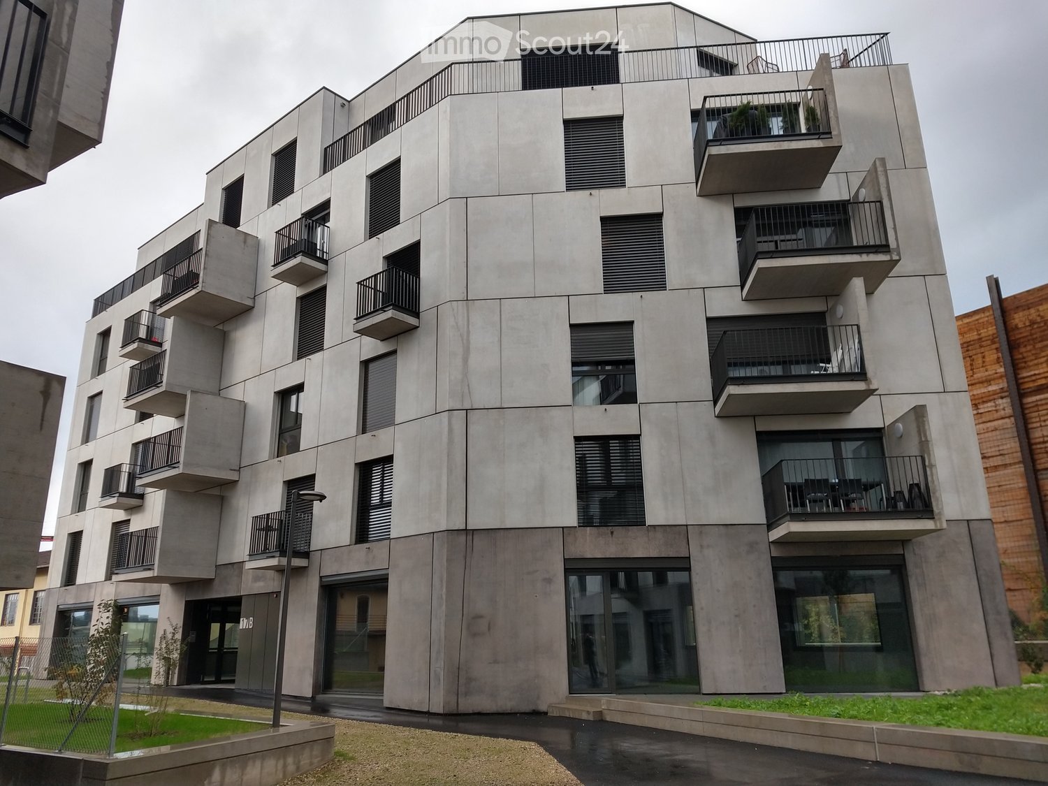 Modern multi-story residential building with concrete facade, multiple balconies, large windows, and a raised ground floor entrance.