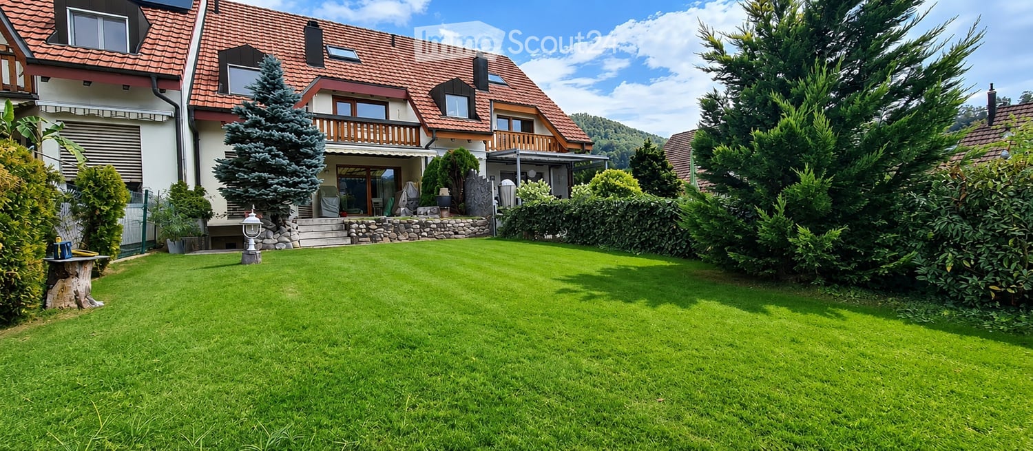 Well-maintained garden with a neatly trimmed lawn, surrounded by trees and shrubs. A two-story house with a red-tiled roof and balconies is visible in the background.