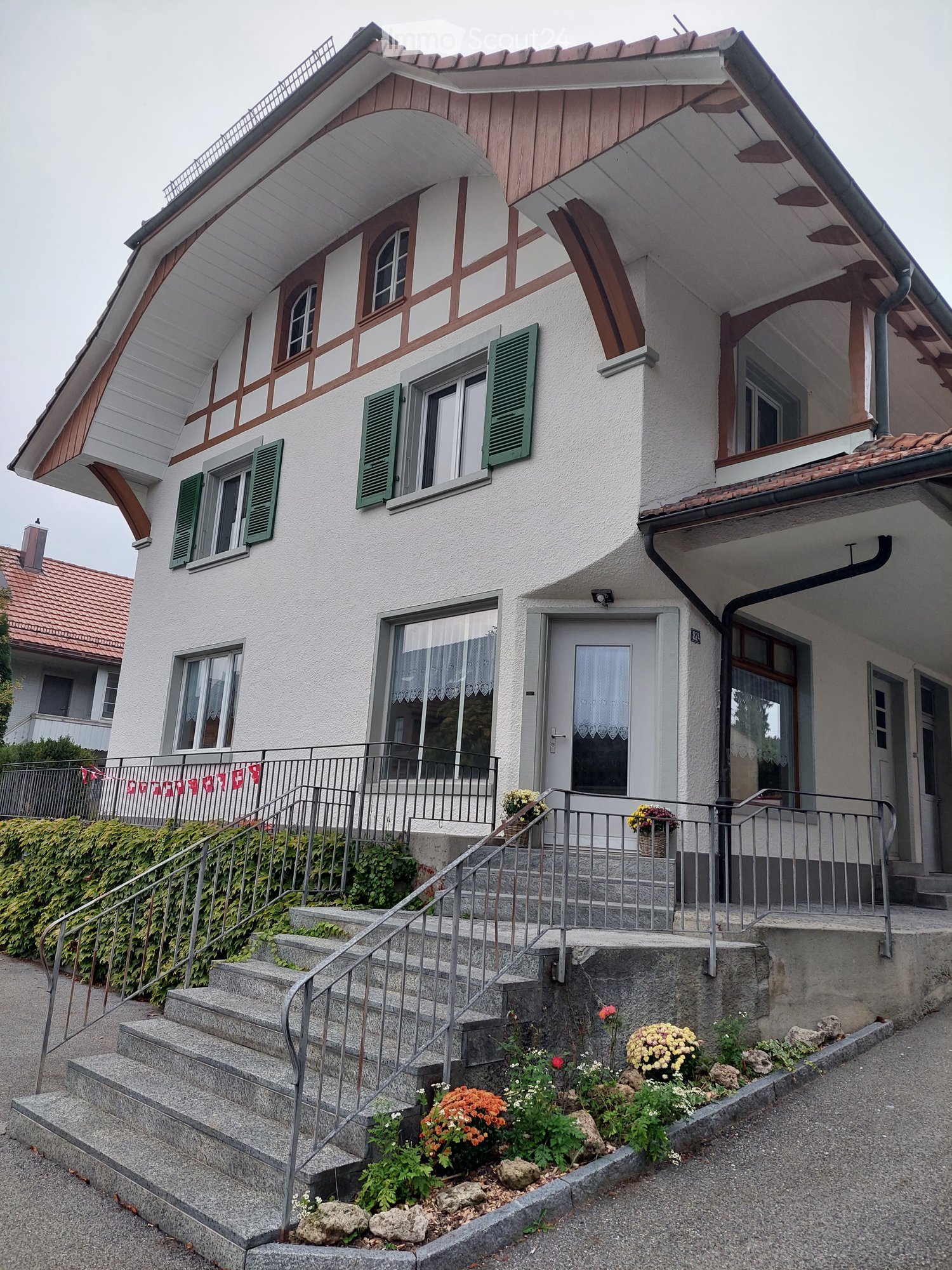 Two-story house with white walls, green shutters, and a wooden roofline. Features a staircase leading to the entrance, surrounded by a small garden with flowers and rocks.