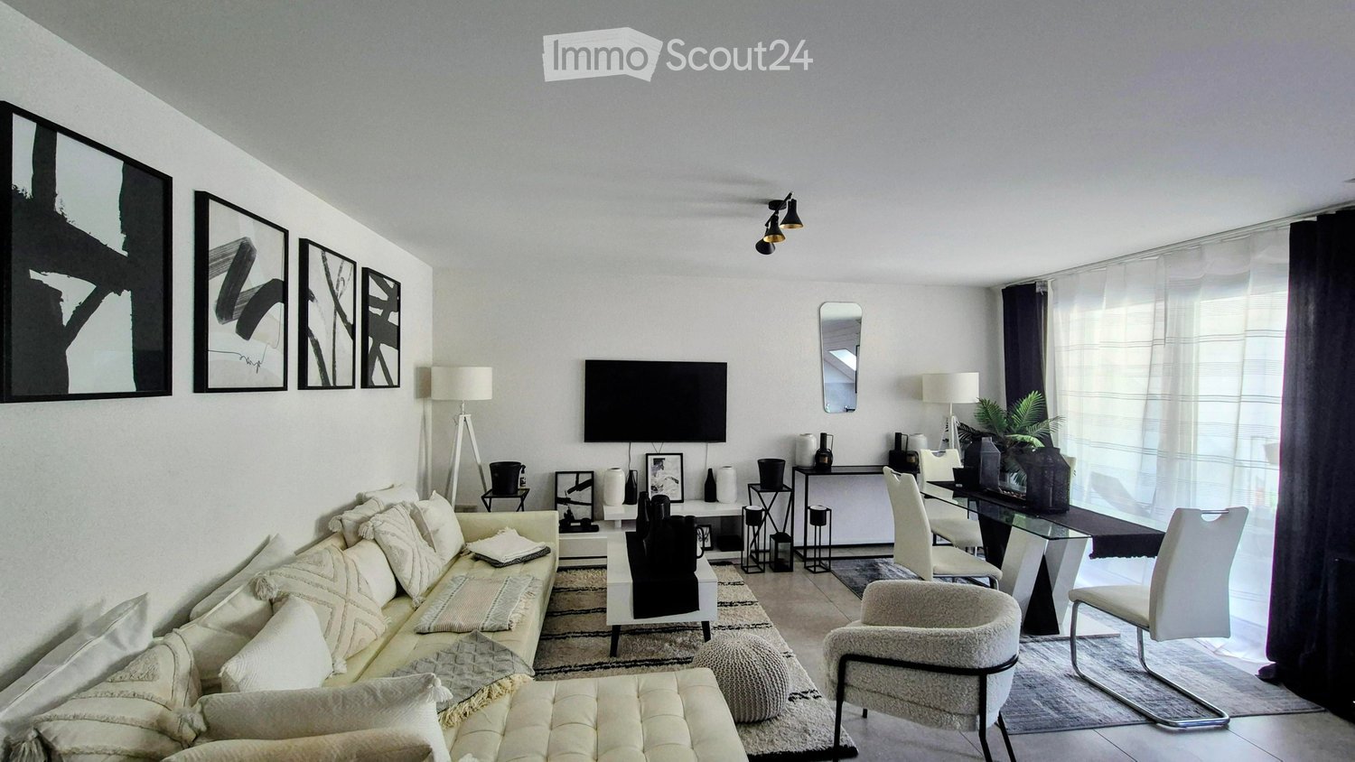 Modern living room with a cream sofa, ottoman, white chairs, glass dining table, television, lamps, and decorative items.
