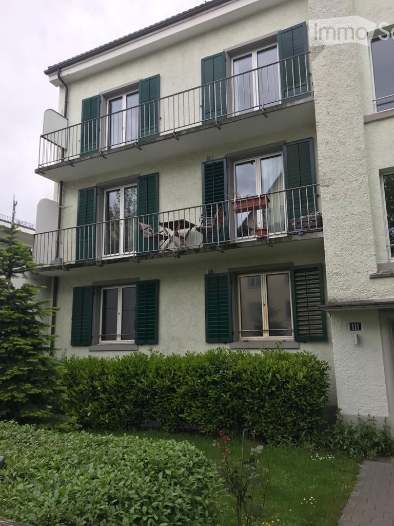 Three-story residential building with white facade, green shutters, and multiple balconies. Balconies feature outdoor furniture and potted plants.