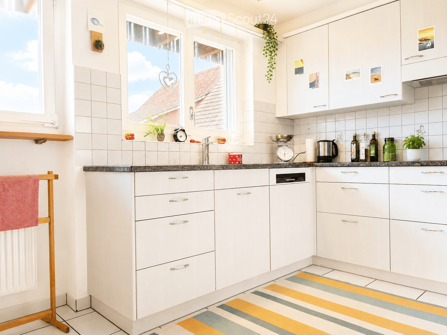 Bright, modern kitchen with white cabinets, tiled walls, and a dark countertop. Features a dishwasher, faucet, potted plants, and decorative items on the counter.