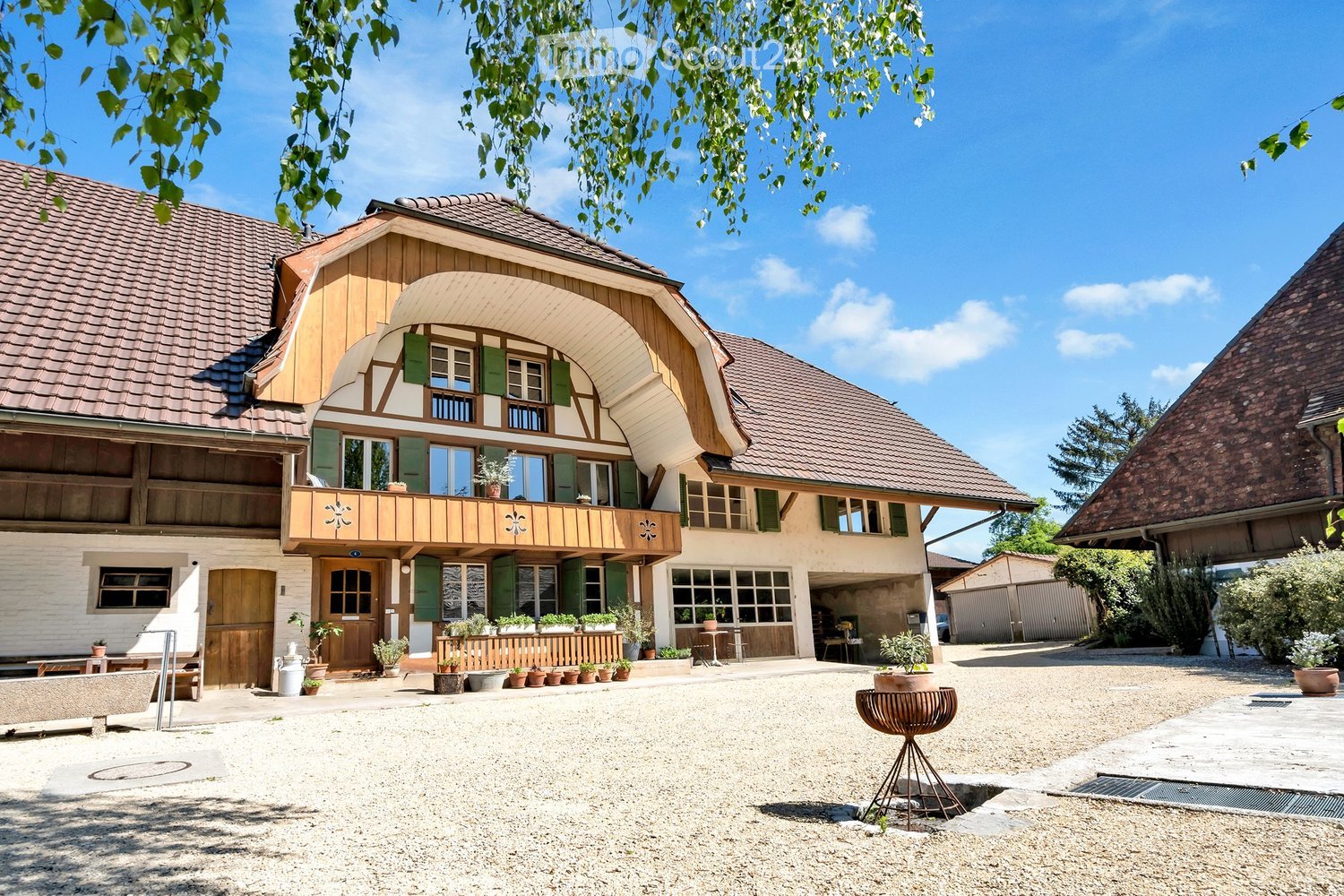 A two-story traditional house with a wooden facade, green shutters, and a covered balcony. The house features multiple windows, a wooden door, and a gravel driveway with potted plants and outdoor furniture.