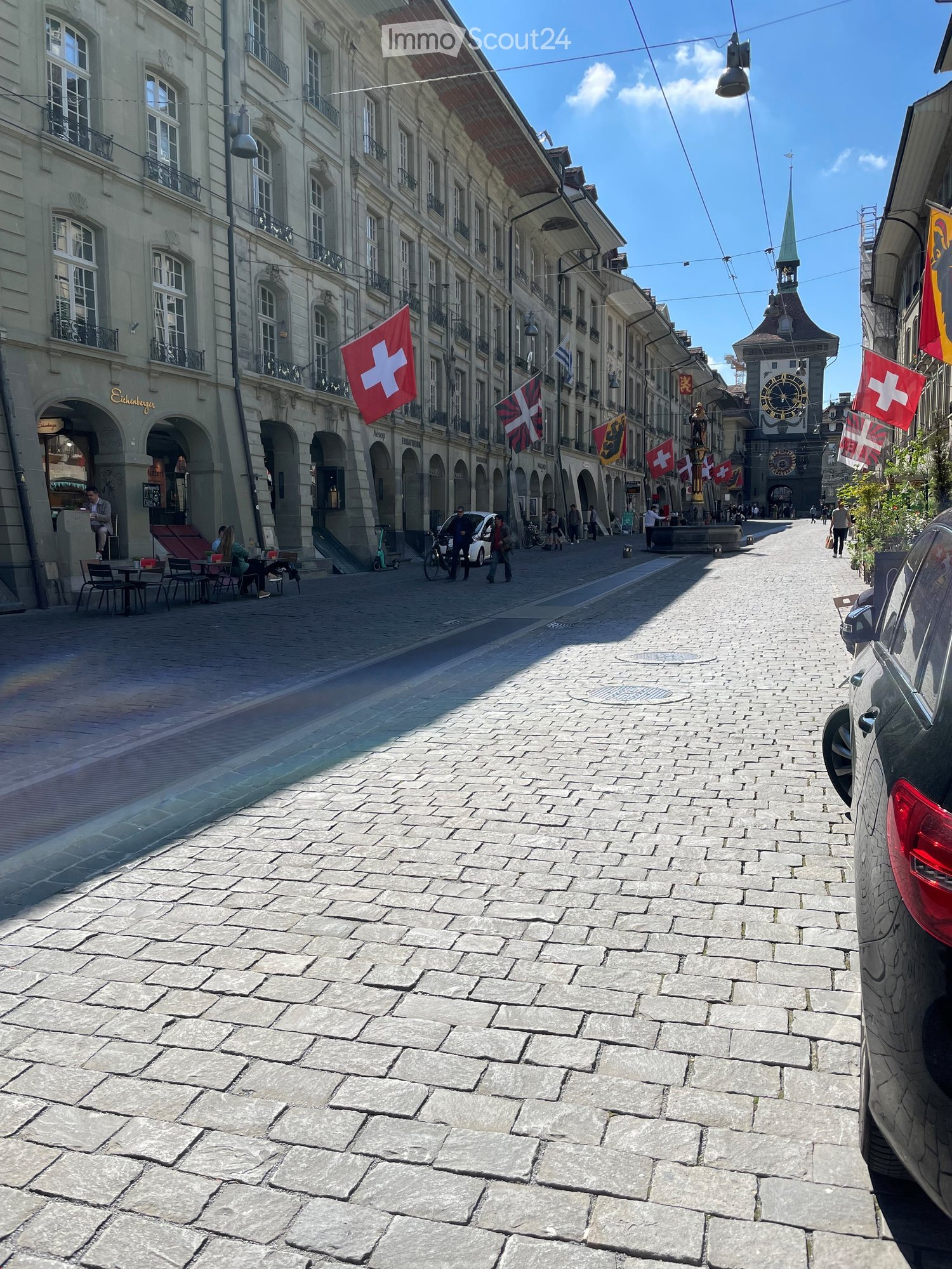 Cobblestone street with buildings on both sides, Swiss flags, people walking, a clock tower, and a car parked on the side.