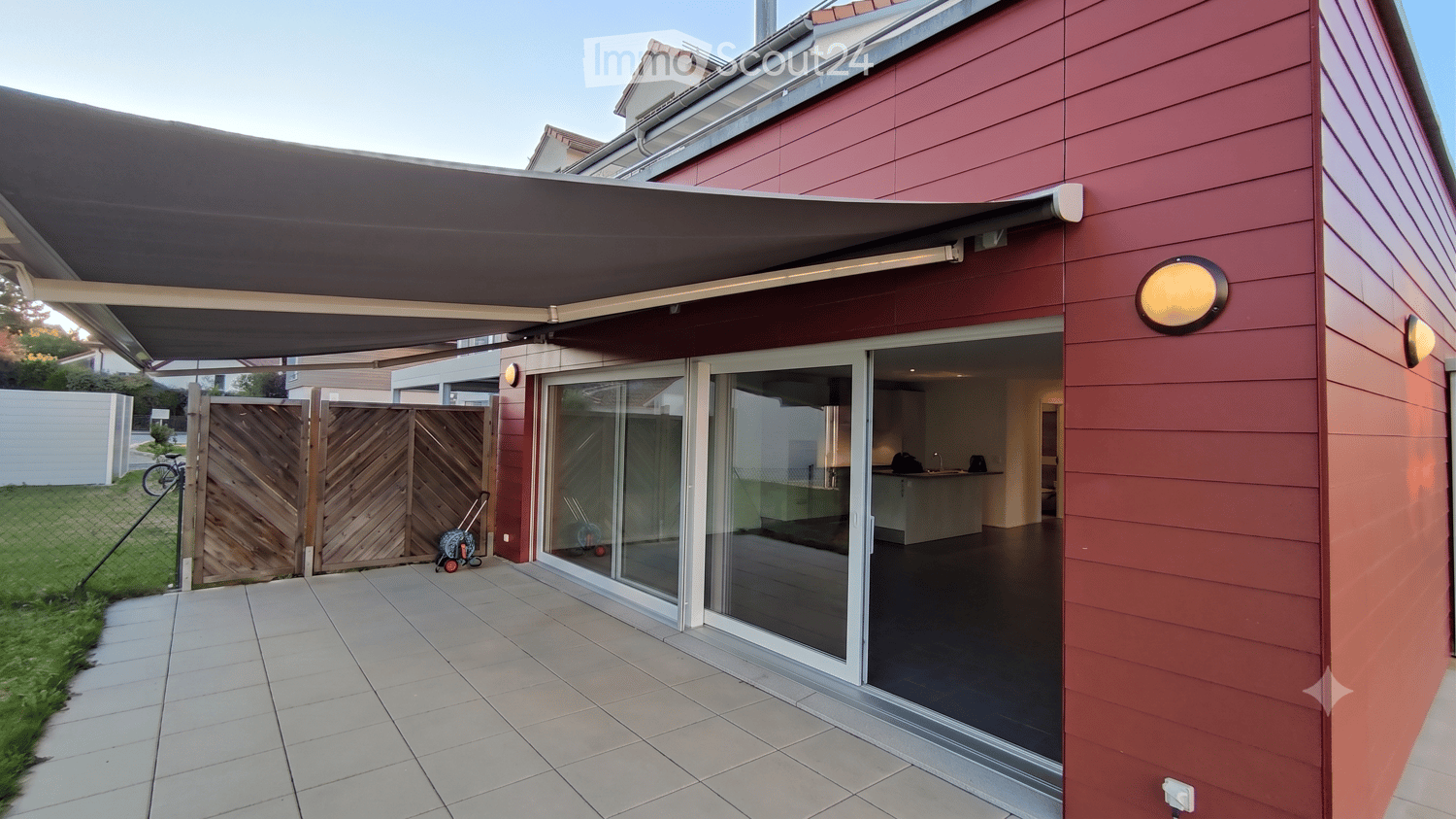 Modern outdoor patio area with a retractable awning, large glass sliding doors leading into a room, tiled floor, wooden gate, and a small grassy area.
