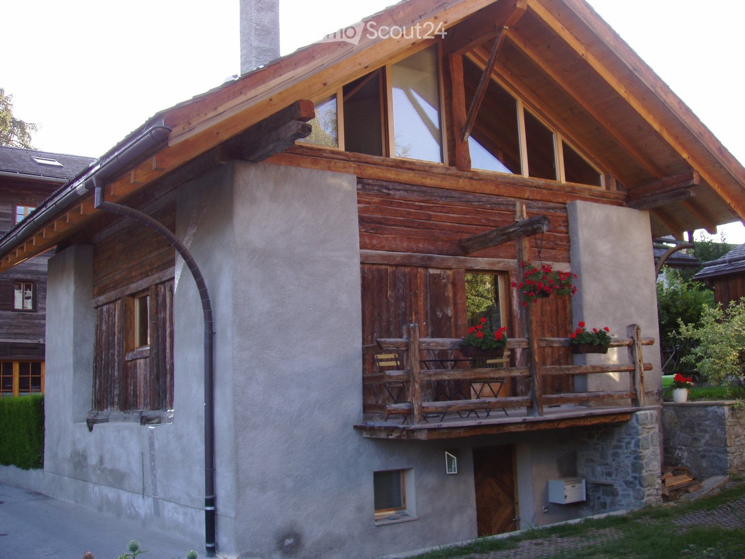 Two-story wooden house with a stone base, large windows, and a balcony with wooden furniture and flower pots.