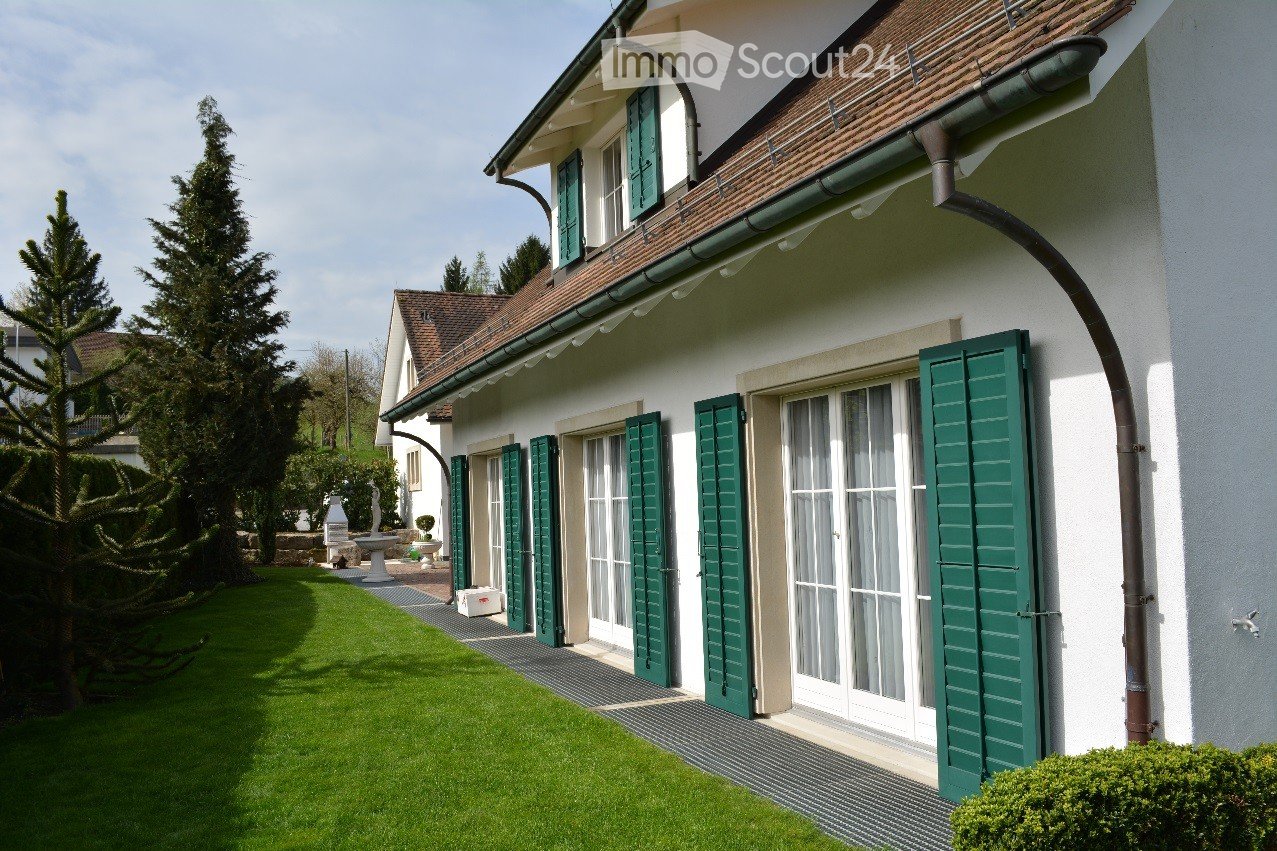 A well-maintained garden area with a neatly trimmed lawn, a large evergreen tree, and a white house with green shutters and a brown tiled roof in the background.