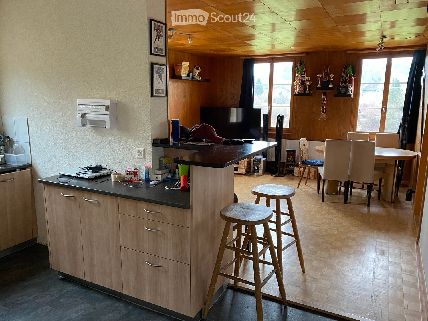 A modern kitchen and dining area with wooden cabinets, a black countertop, a laptop, a TV, and a dining table with chairs.