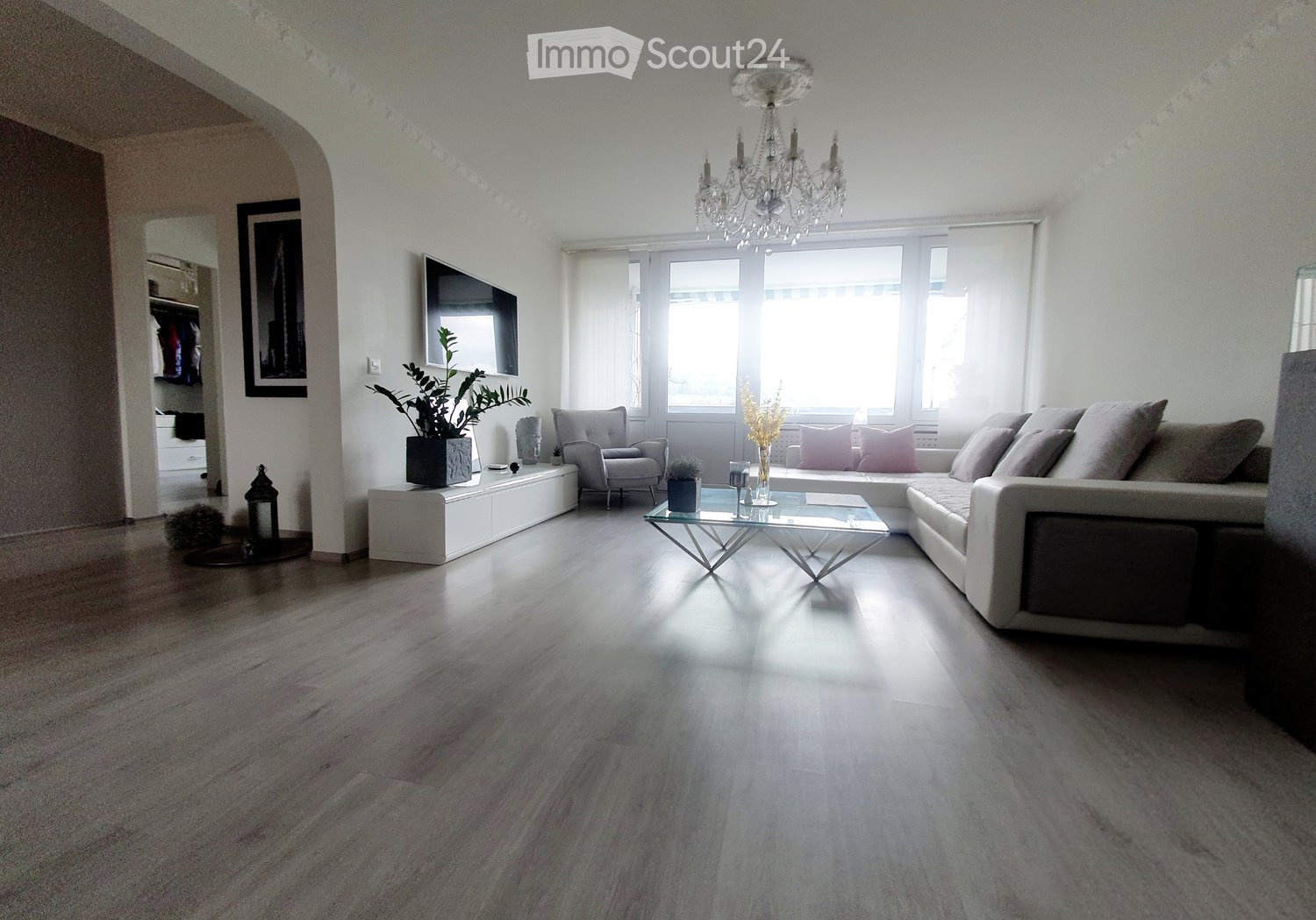 Modern living room with light-colored wooden flooring, a large sectional sofa, a glass coffee table, an armchair, a chandelier, a TV, and decorative plants.