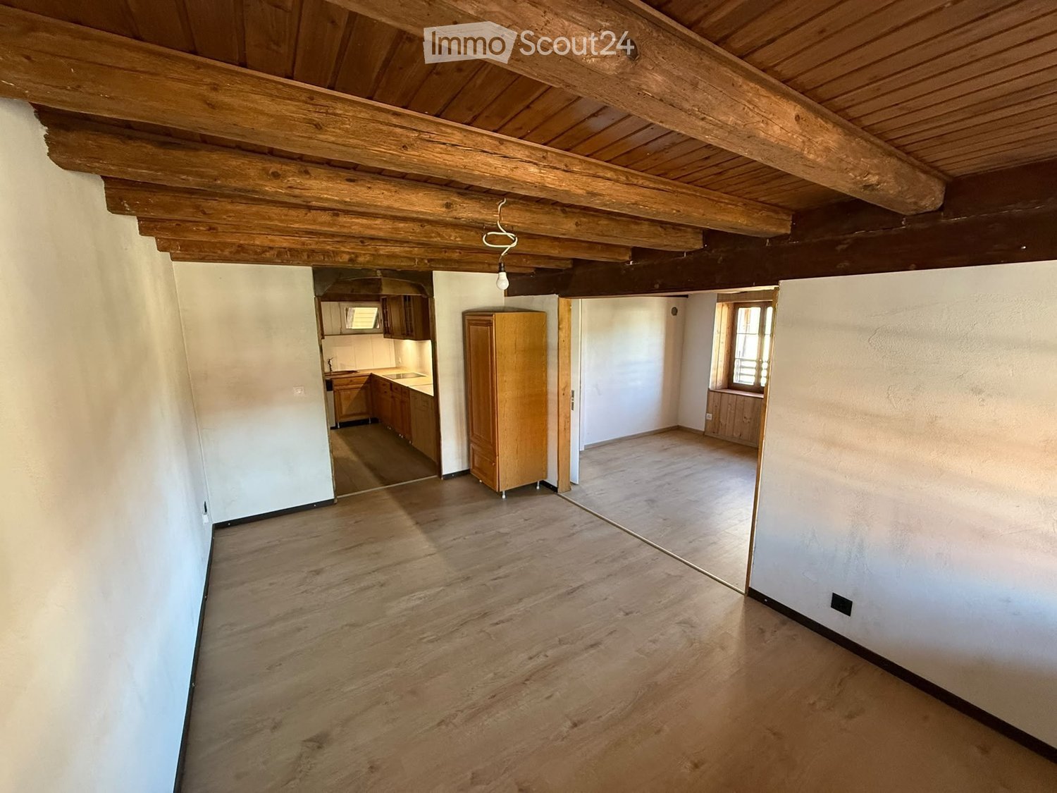 Empty room with wooden floor and exposed wooden ceiling beams. Adjacent kitchen area visible with cabinets and appliances.