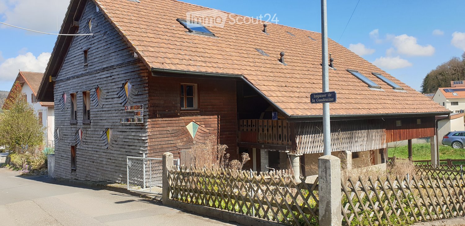 Traditional two-story house with a red-tiled roof, wooden facade, and decorative elements. Features include a balcony, small garden area, and a wooden fence.