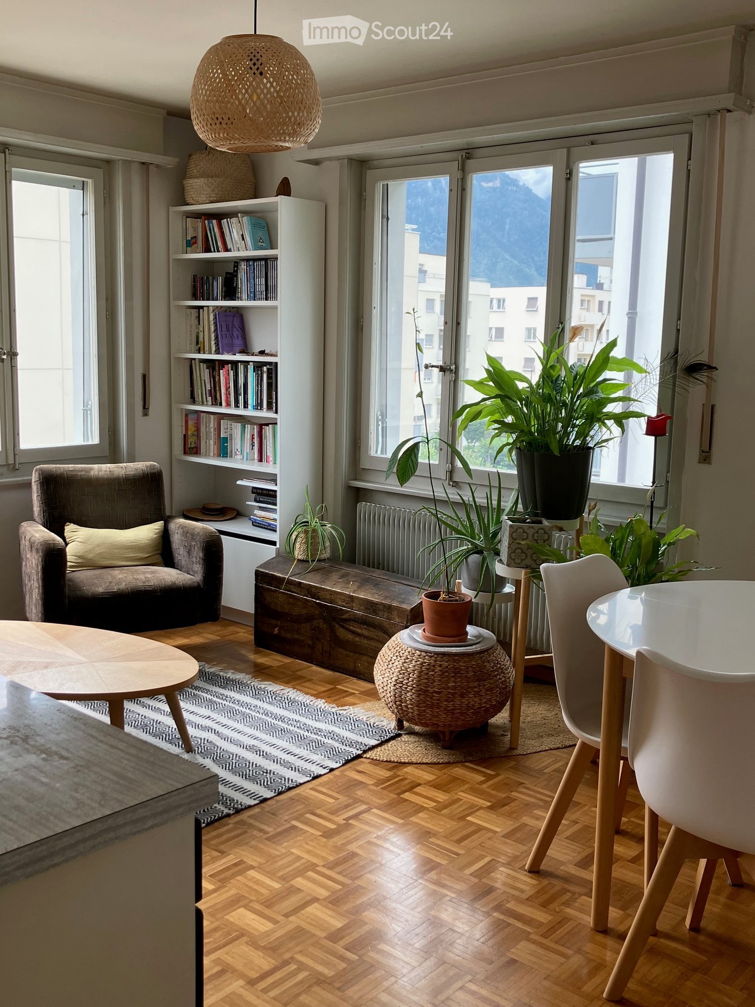 Living room with wooden flooring, large windows offering a mountain view, a sofa, a coffee table, a bookshelf, and several potted plants.