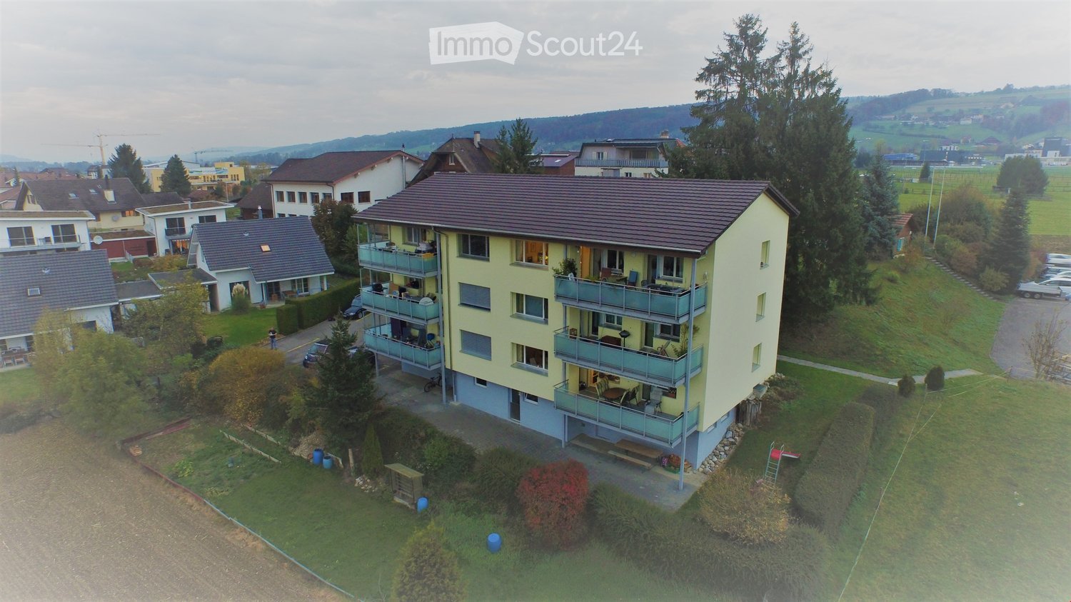 Aerial view of a multi-story residential building with balconies, surrounded by greenery and neighboring houses.