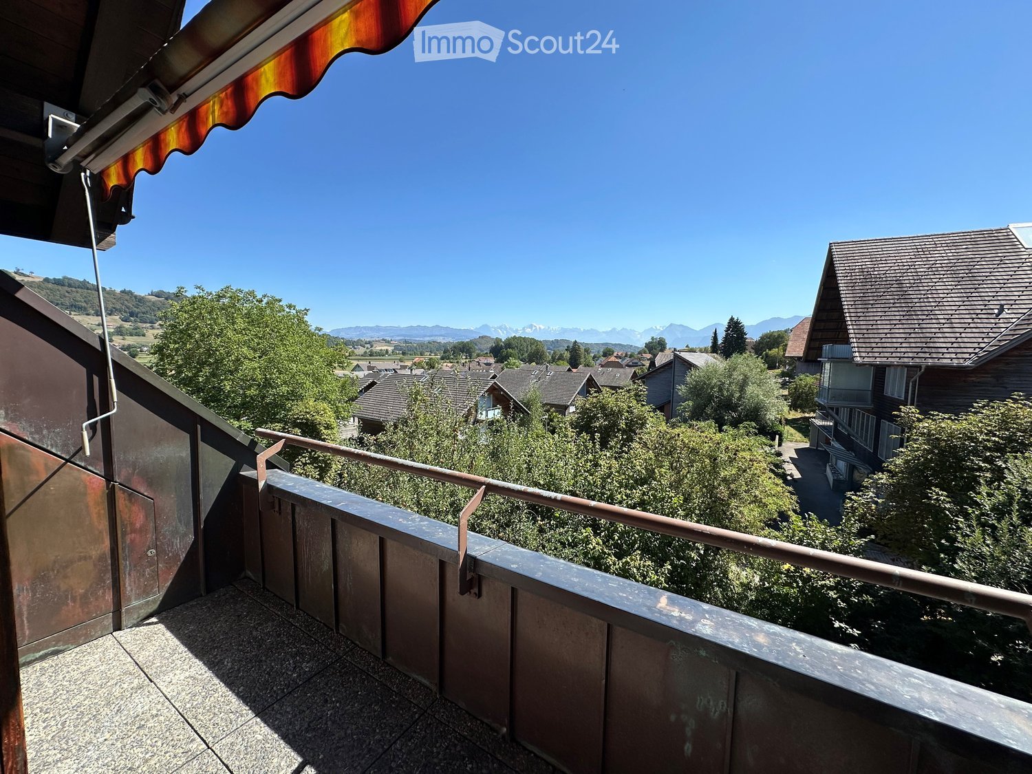 Balcony with metal railing, overlooking a residential area with houses, trees, and distant mountains under a clear blue sky.