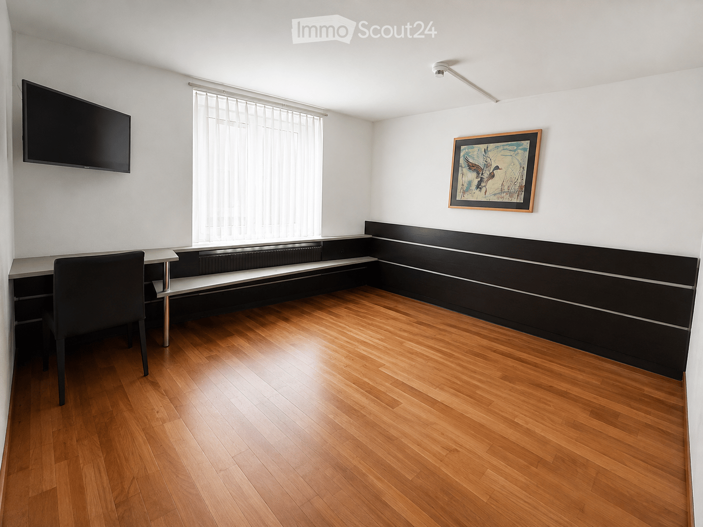 A modern, minimalist room with wooden flooring, a black chair, a small table, a mounted screen, and a framed picture on the wall.