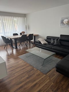 Bright, modern living and dining area with hardwood flooring, large windows with sheer curtains, a black sectional sofa, a glass coffee table, a dining table with six chairs, a floor lamp, and a decorative picture frame on the wall.