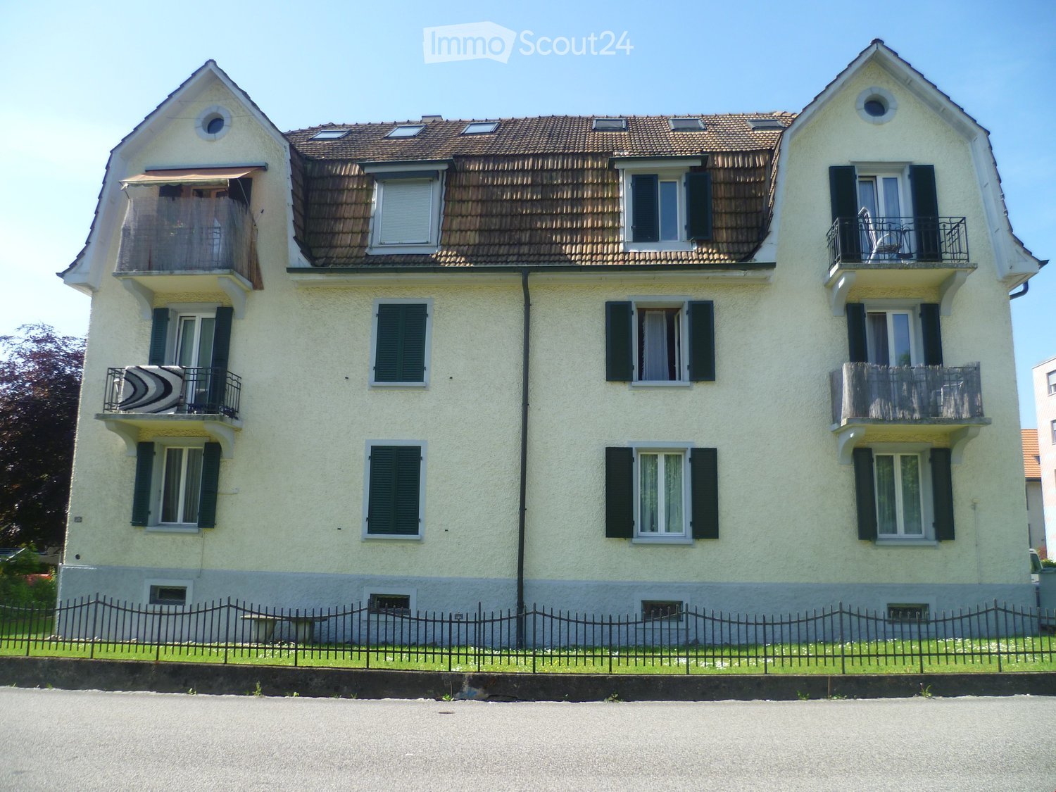 Two-story house with light yellow walls, dark green shutters, multiple windows, and balconies on both floors.