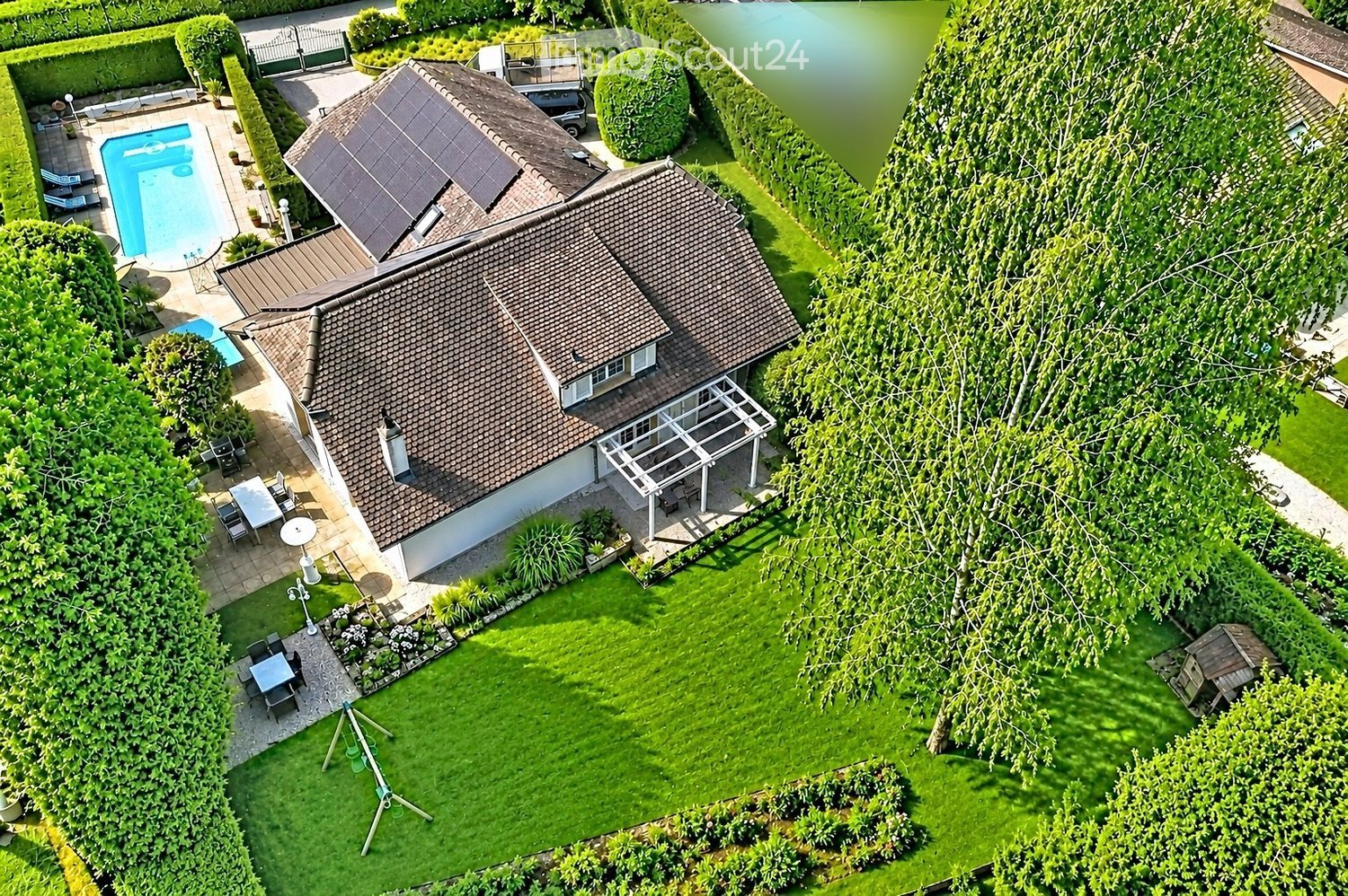Aerial view of a house with a well-maintained garden, swimming pool, patio furniture, and a covered outdoor seating area.